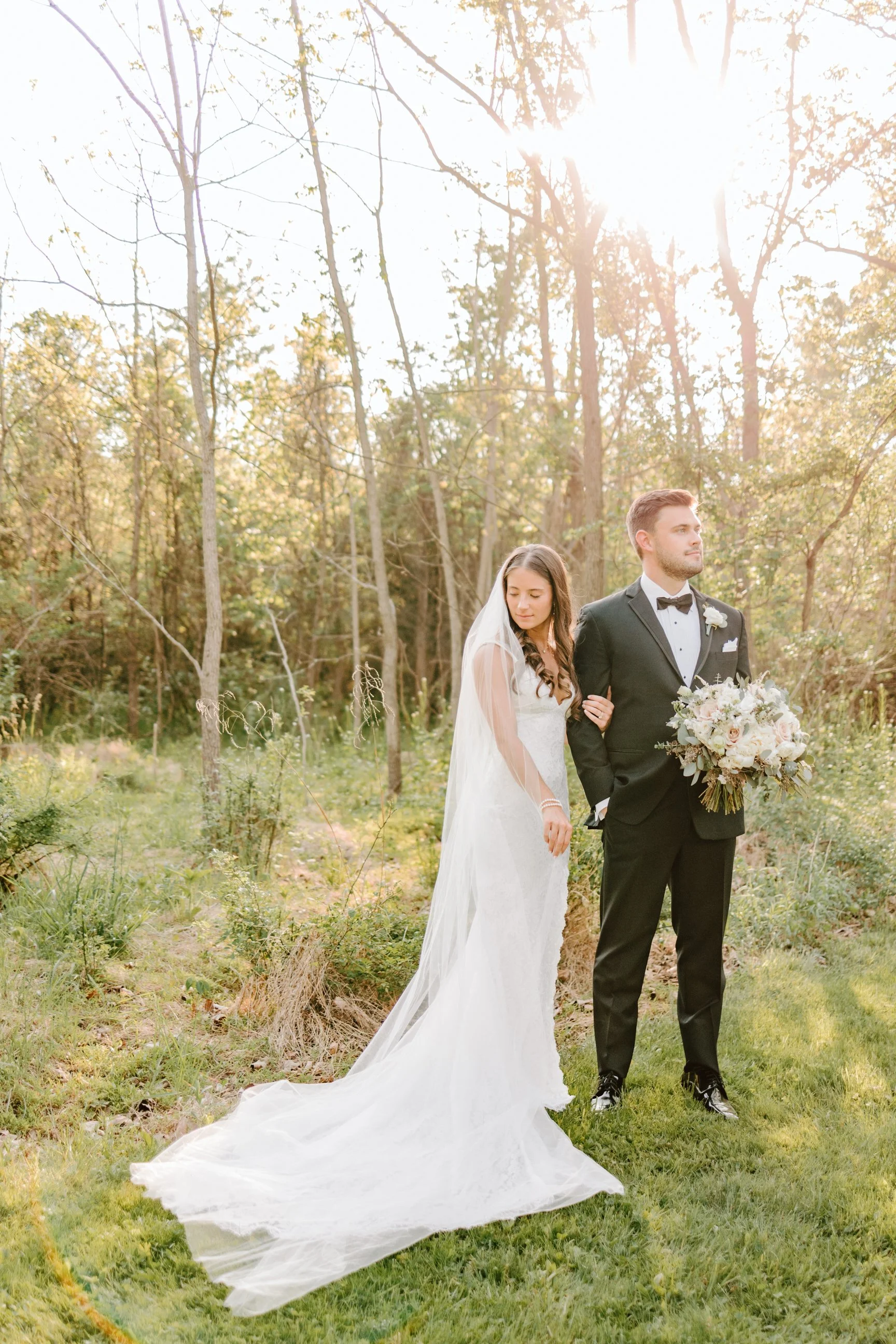 A bride and groom stand outdoors in a wooded area during sunset, with the bride in a white wedding gown and veil holding onto the groom's arm, who is in a black tuxedo holding a bouquet of flowers.