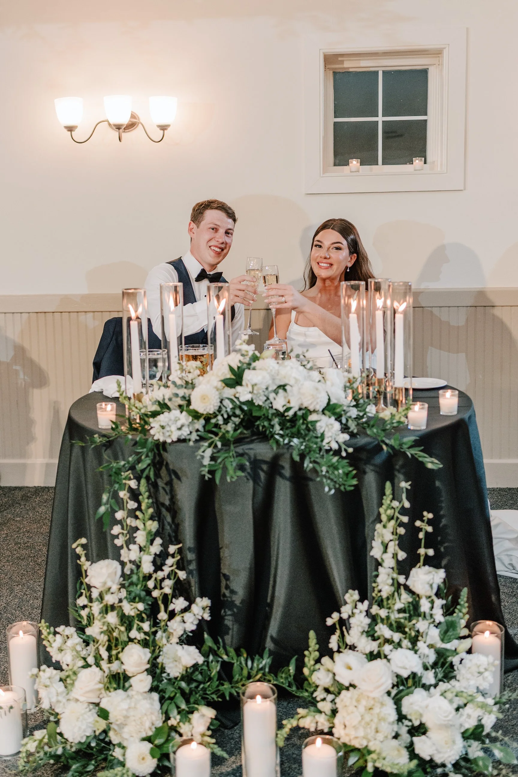 A newlywed couple sitting at a wedding reception table, raising champagne glasses for a toast, with a floral centerpiece and candles.