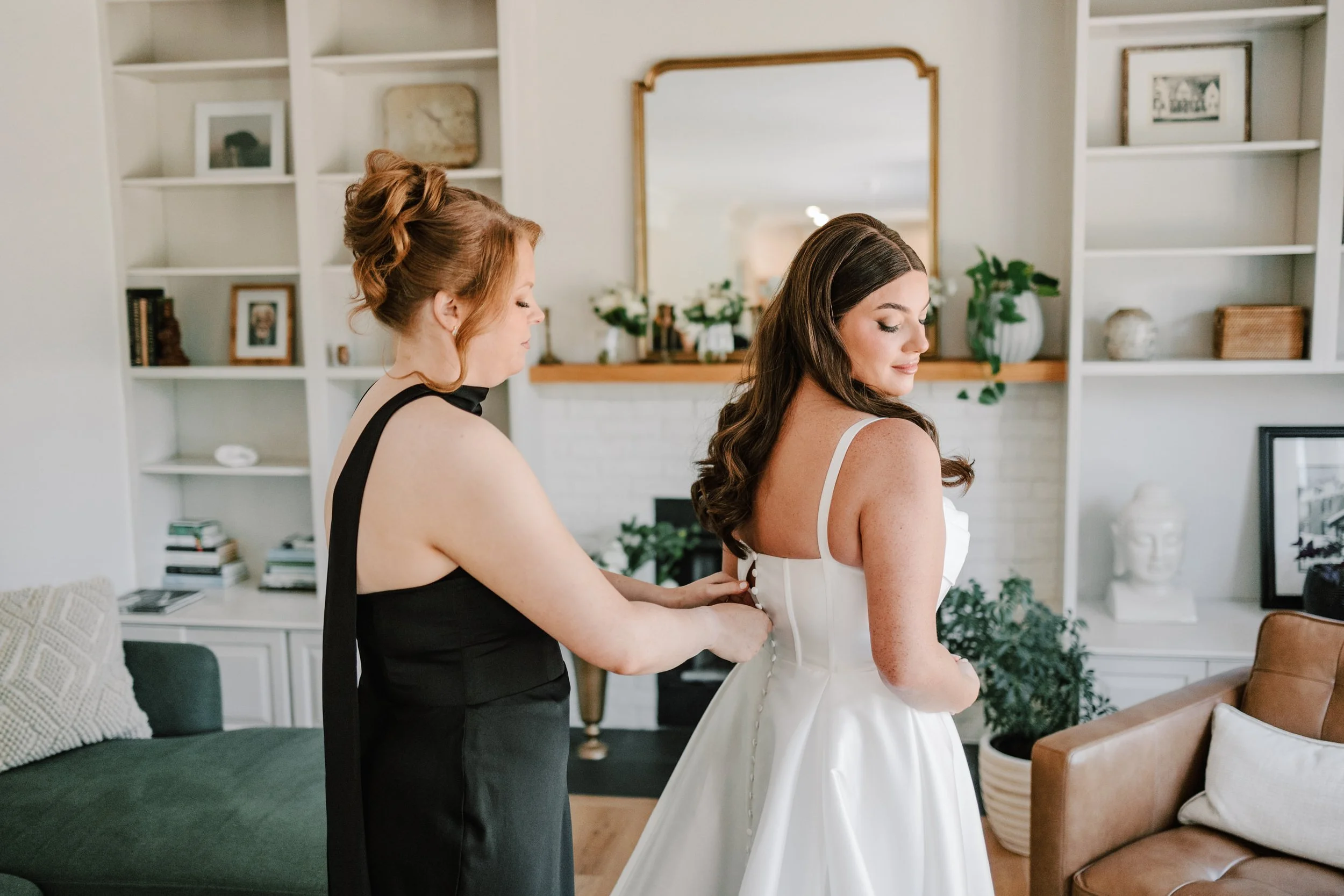 A woman in a white wedding dress is being helped into her dress by another woman in a black dress in a living room.