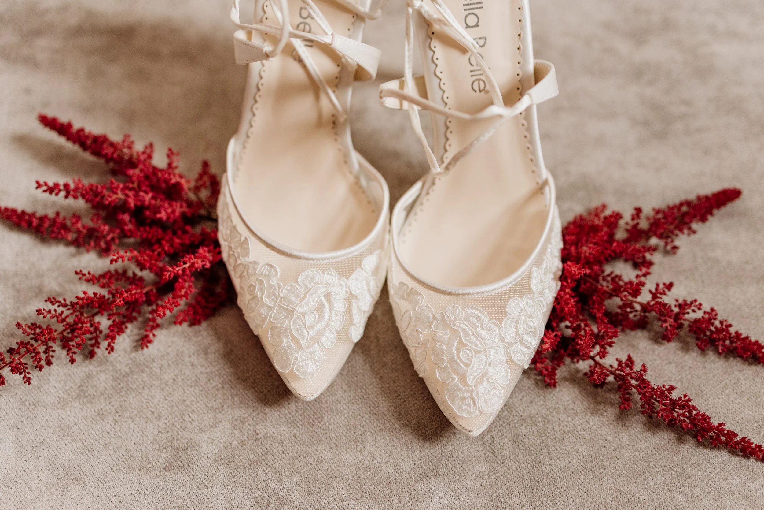White lace bridal shoes placed on a beige surface with red decorative artificial flowers.