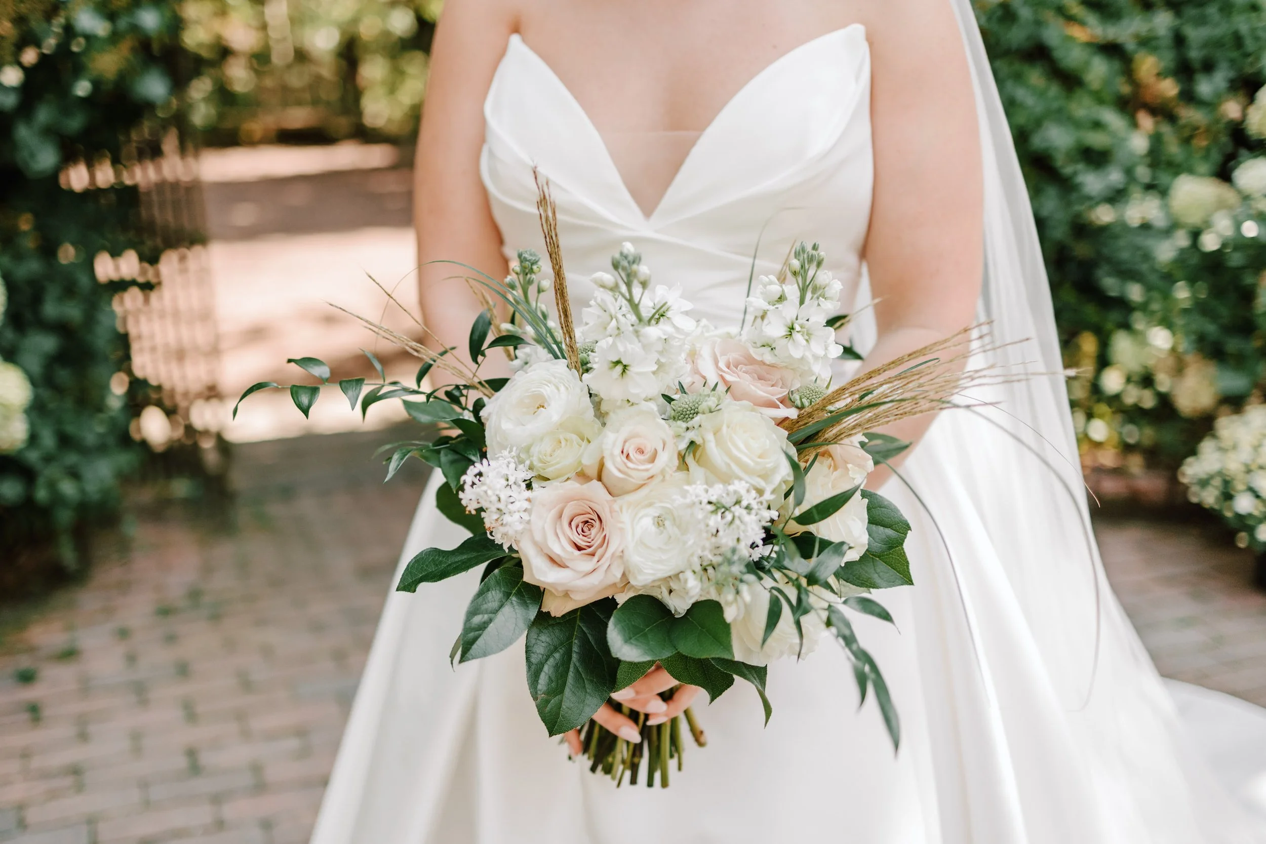 A bride holding a bouquet of white and blush pink roses, white flowers, greenery, and decorative grasses in an outdoor garden setting.