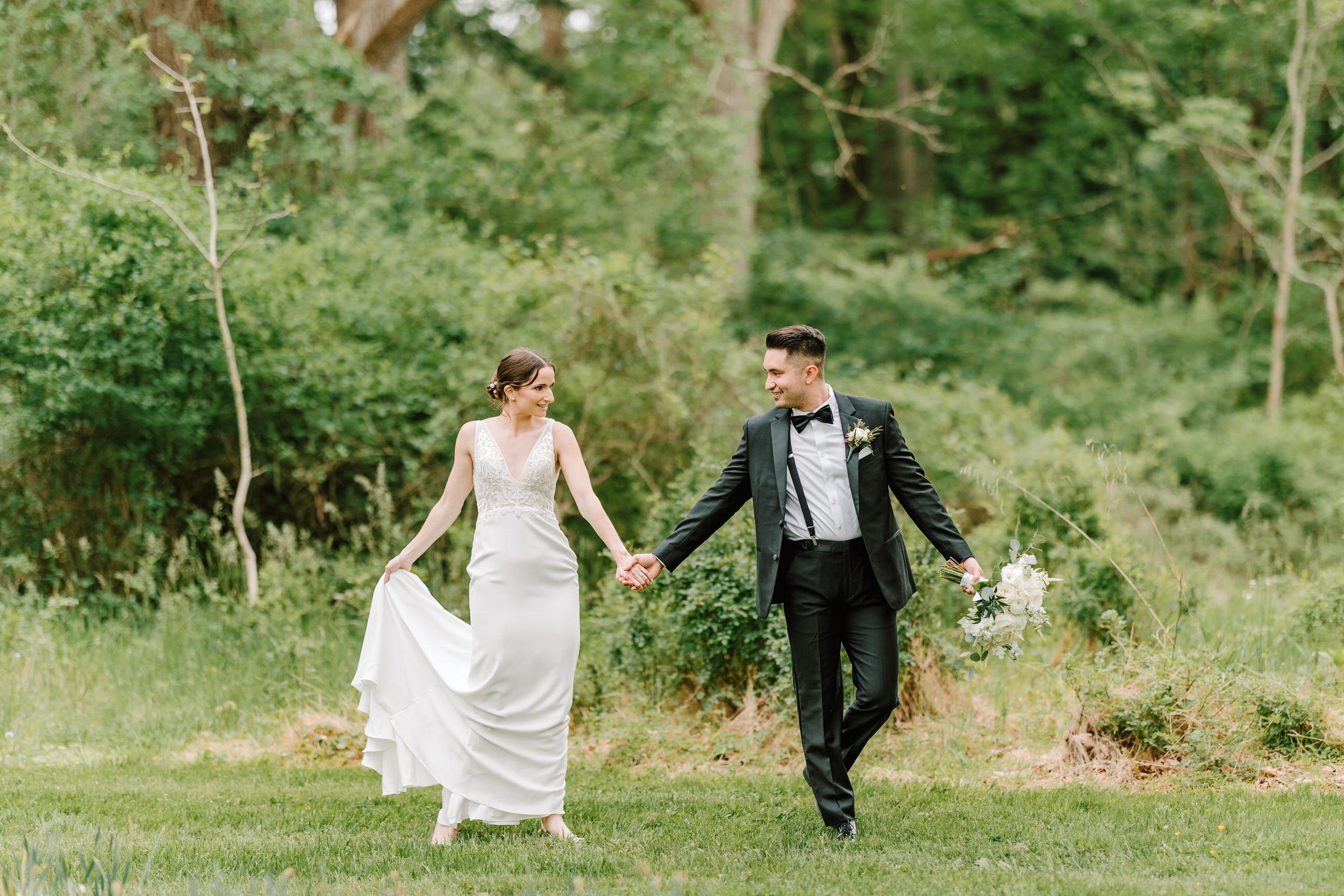 A bride and groom holding hands and smiling at each other outdoors in a lush green park, with the groom holding a bouquet of flowers.