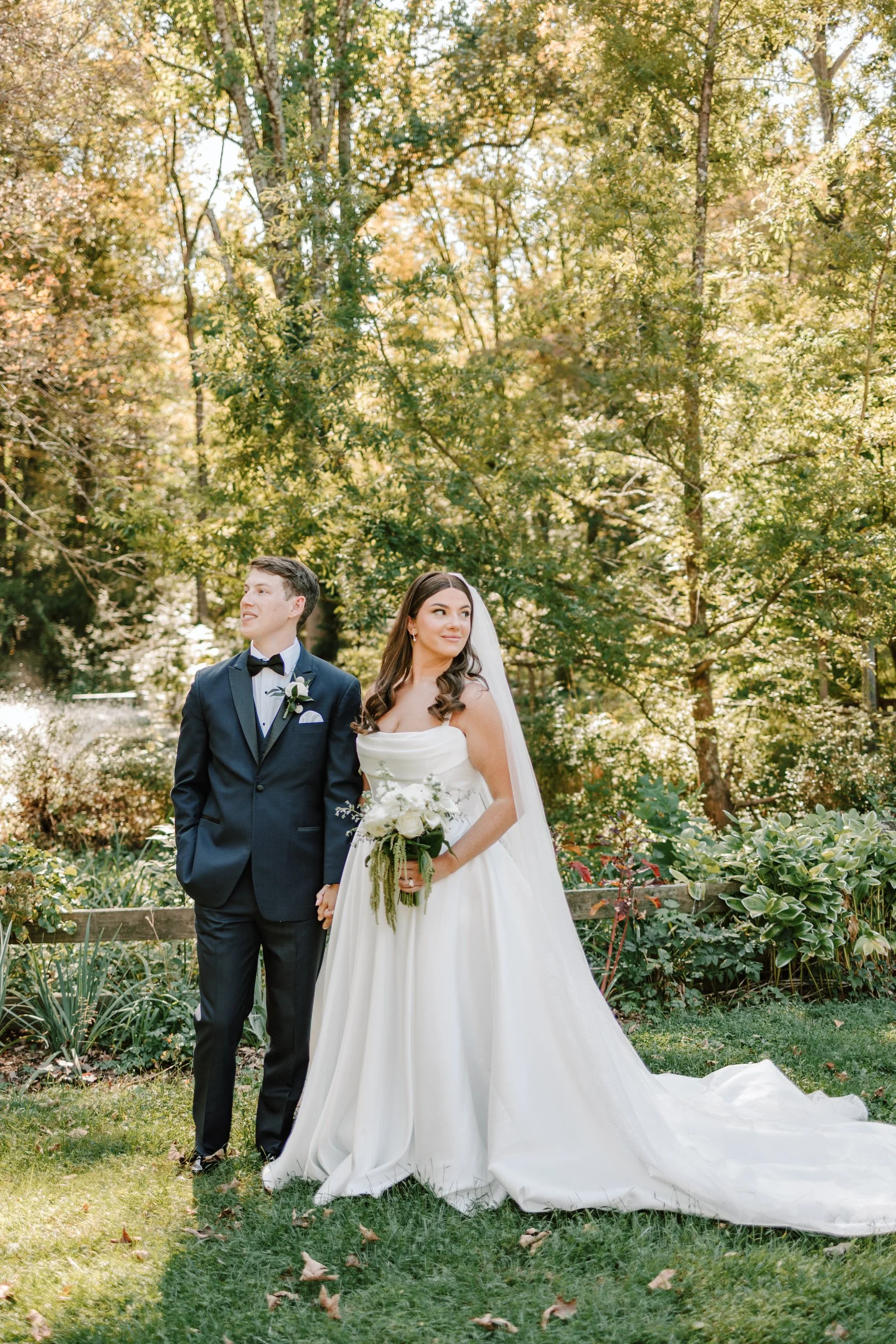 A bride and groom standing outdoors in a garden with trees and green foliage. The bride is wearing a white wedding gown and holding a bouquet, while the groom is wearing a navy blue suit with a bow tie. They are holding hands and appear to be posing 
