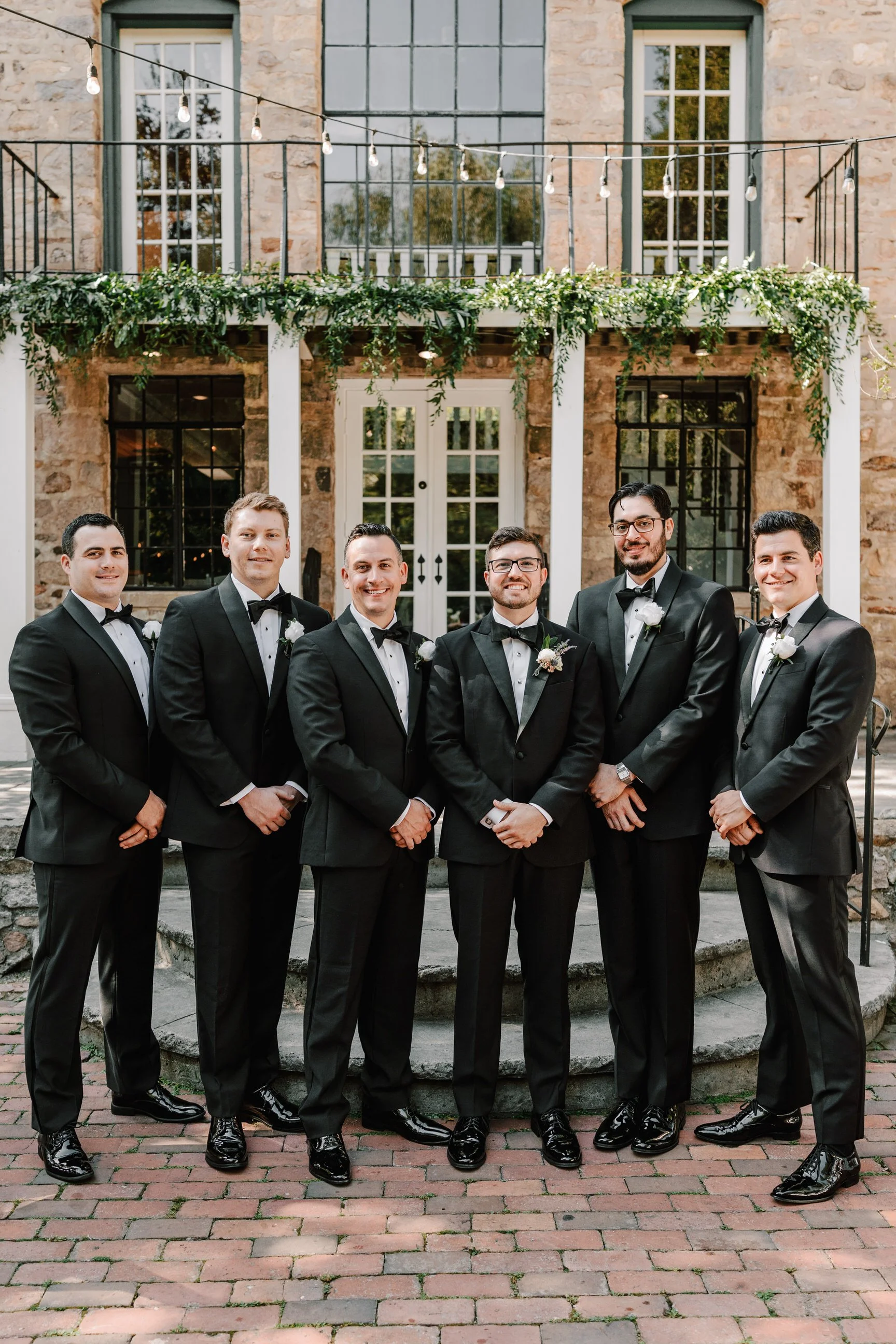 Group of seven men dressed in tuxedos standing outdoors in front of a stone building with a wooden door and windows, decorated with greenery and string lights.