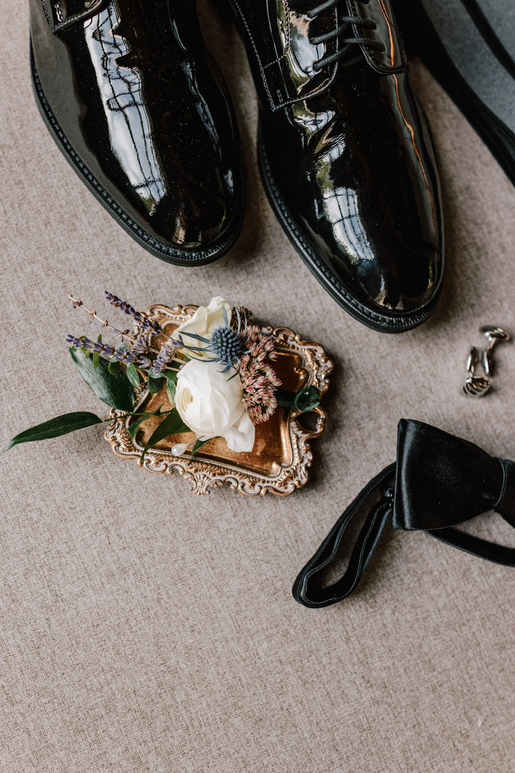Pair of black shiny dress shoes, a small decorative tray with a floral boutonniere, a black bow tie, and a wedding ring on a beige fabric surface.