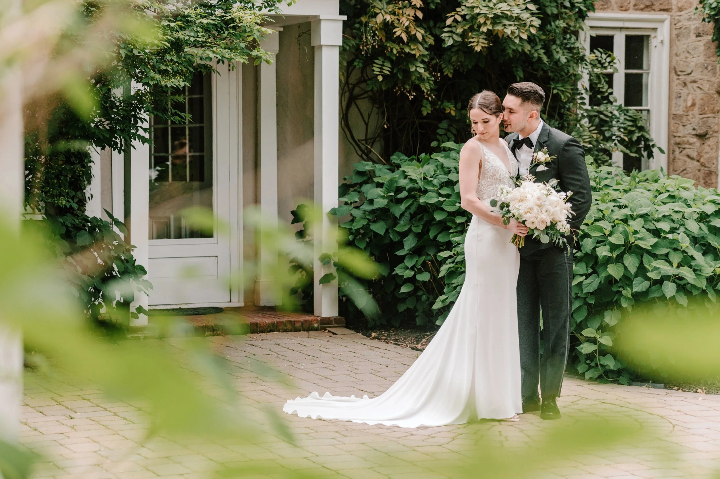 A bride and groom standing close together outside on a brick pathway, the bride holding a bouquet of white flowers, surrounded by green foliage and a white door with windows in the background.