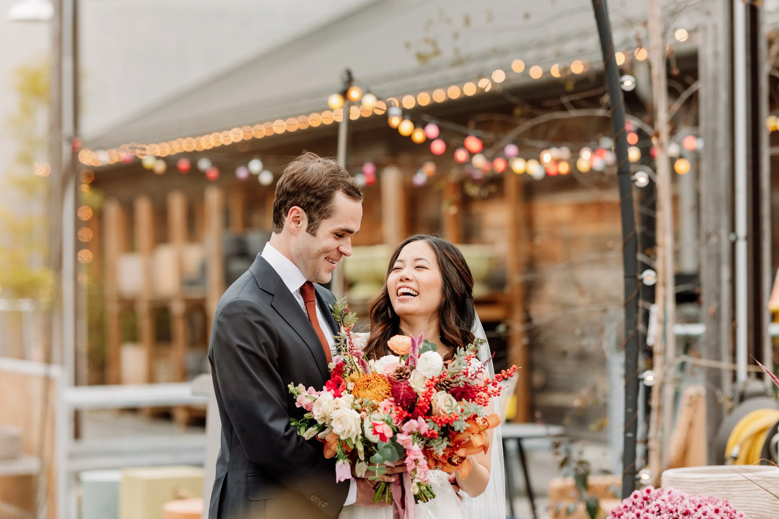 A happy couple at a wedding, with the groom in a black suit and the bride in a white dress holding a colorful bouquet, smiling at each other outdoors decorated with string lights.