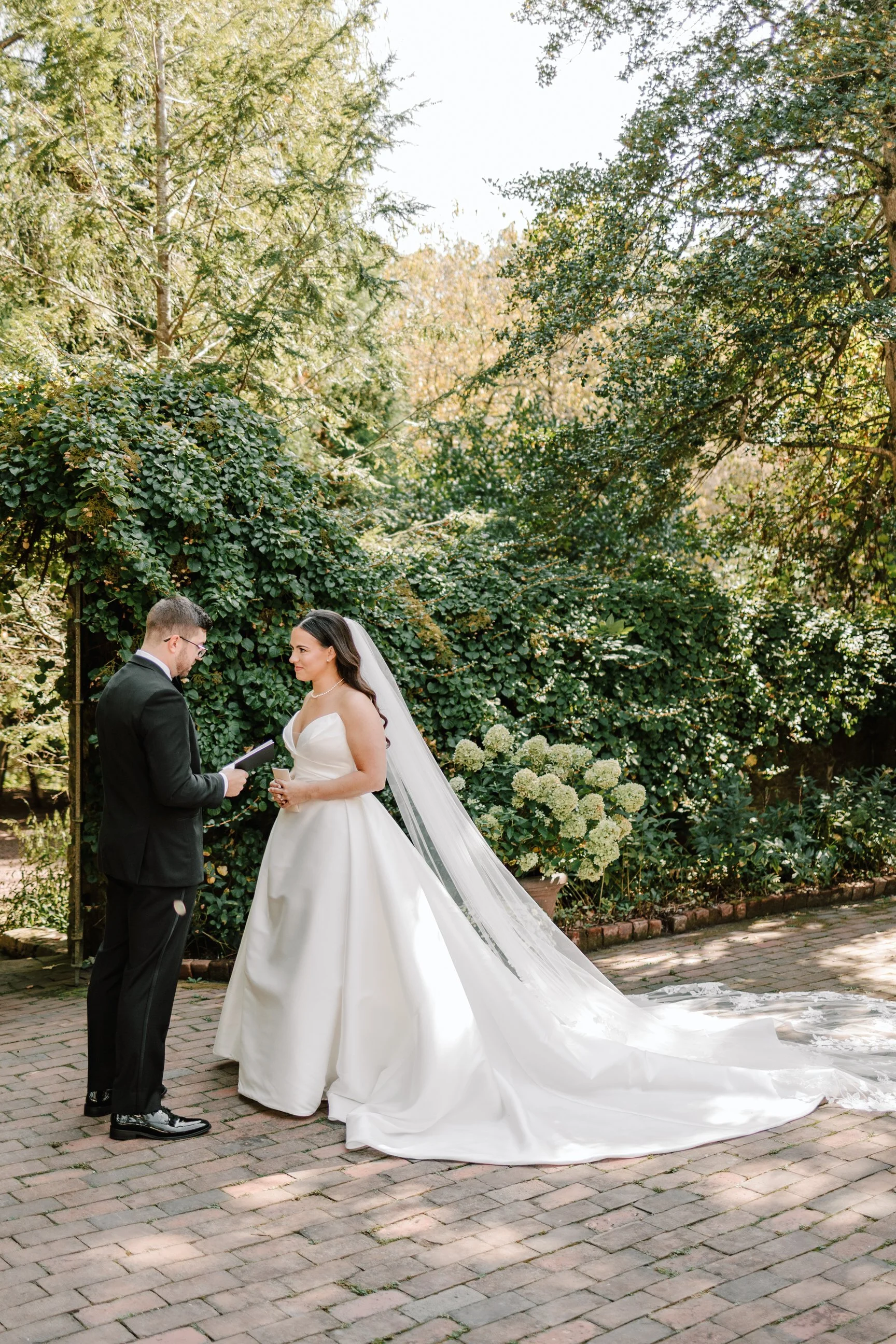 A bride and groom stand facing each other outdoors surrounded by green trees and bushes, with the bride holding a glass and the groom reading from a book or paper during their wedding ceremony.