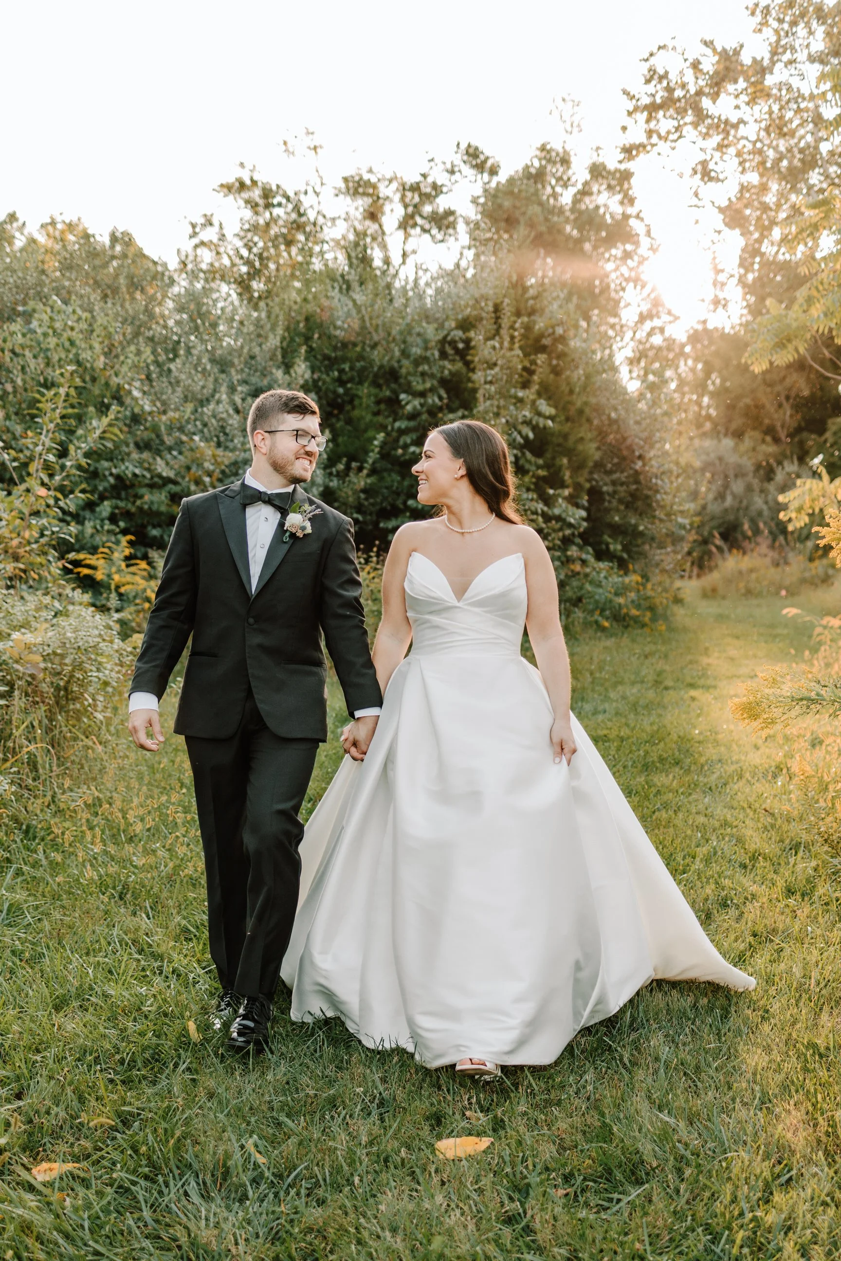 A newlywed couple walking hand-in-hand outdoors in a grassy area with trees, during sunset. The groom is wearing a black tuxedo with a bow tie and glasses, and the bride is wearing a white wedding gown with a sweetheart neckline.