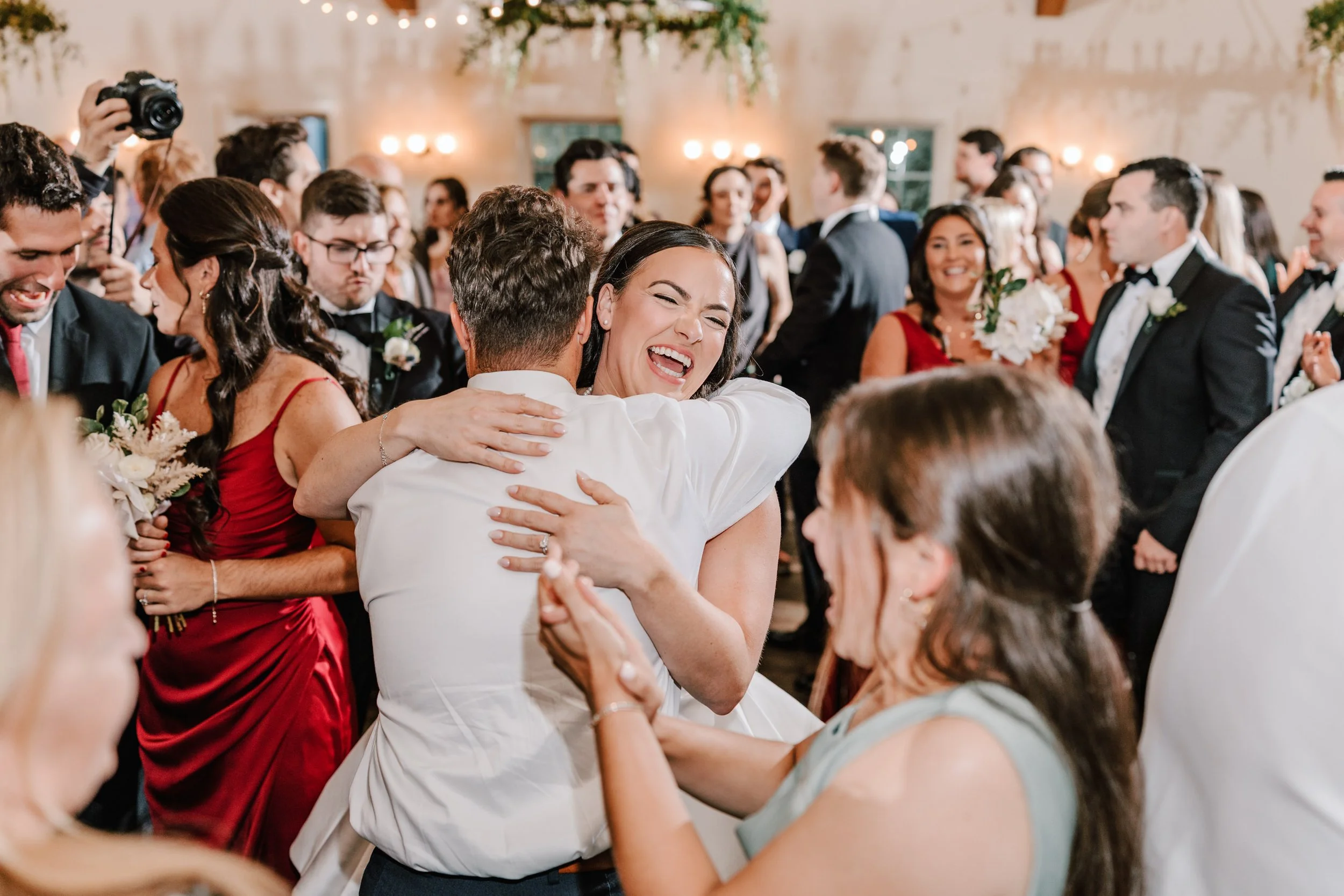 A joyful woman hugging a man at a wedding reception with many guests in formal attire in the background.