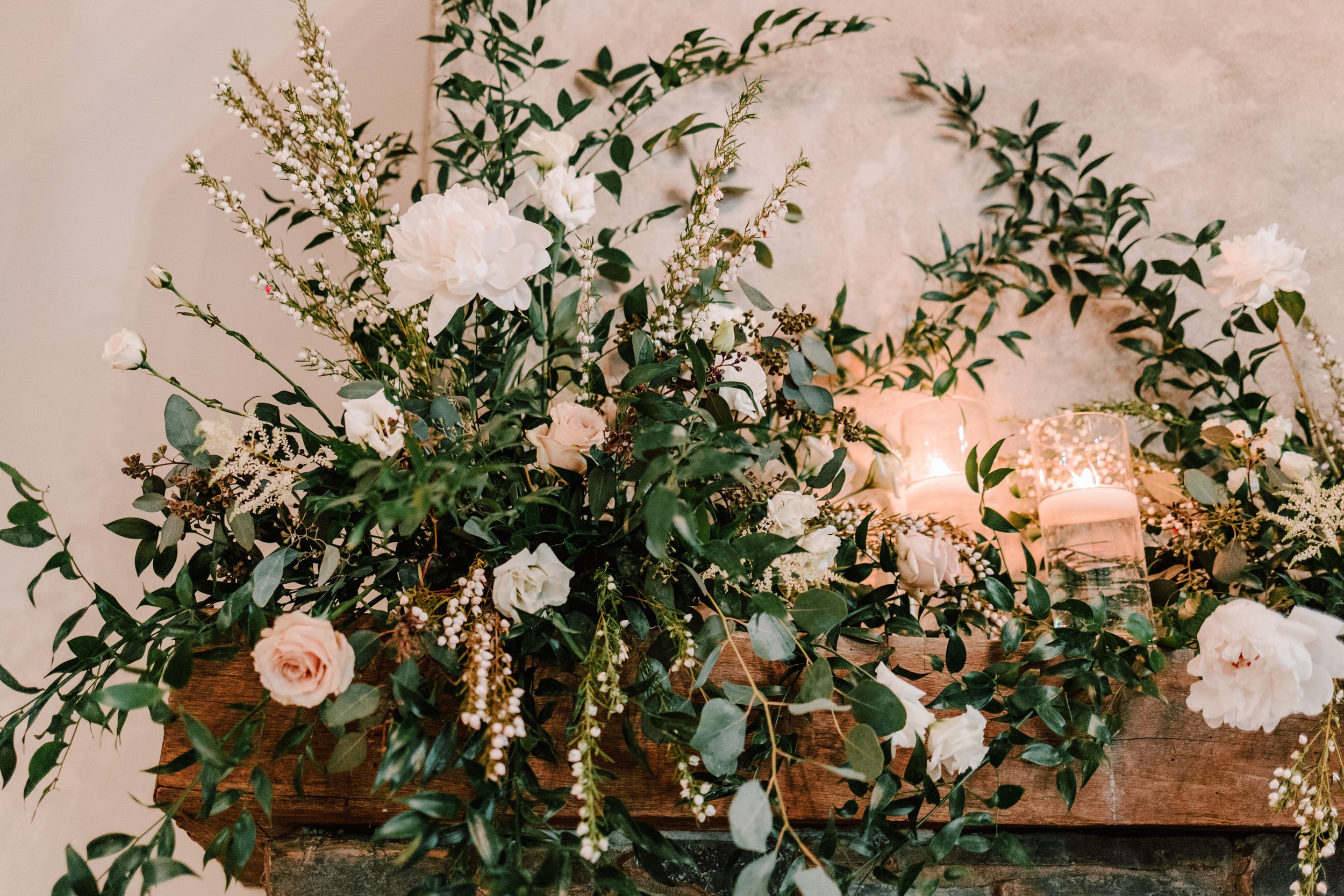 A floral arrangement with white and blush roses, greenery, and small white flowers, illuminated by soft candlelight on a wooden surface against a textured beige wall.