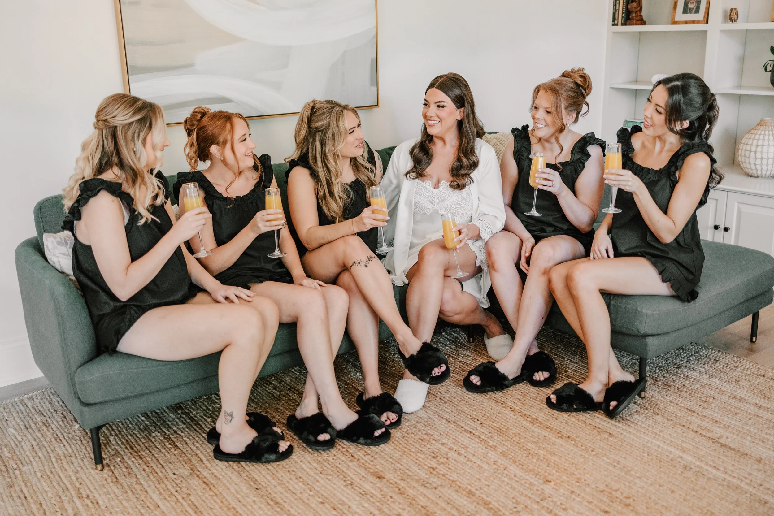 Group of six women sitting on a green couch, holding glasses of orange drinks, smiling and chatting, in a bright living room.