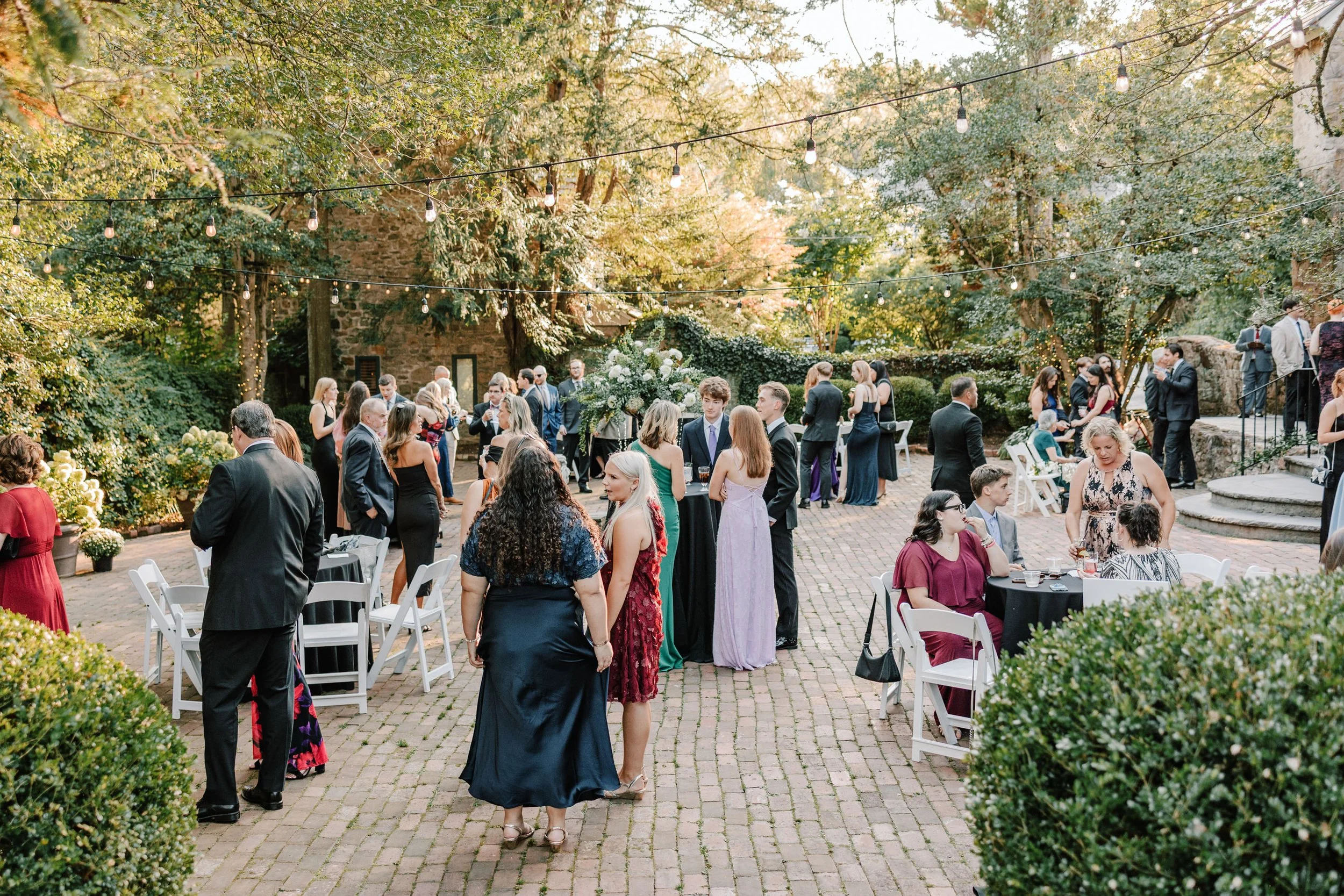 People gathered at an outdoor wedding reception, mingling on a brick patio decorated with string lights and surrounded by greenery and trees.