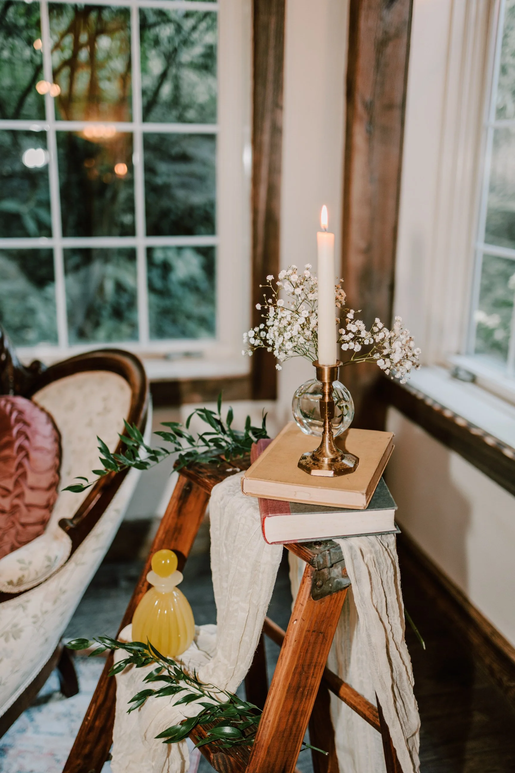 Interior scene with a candle, books, and decorative items on a wooden step ladder by a window with greenery outside.