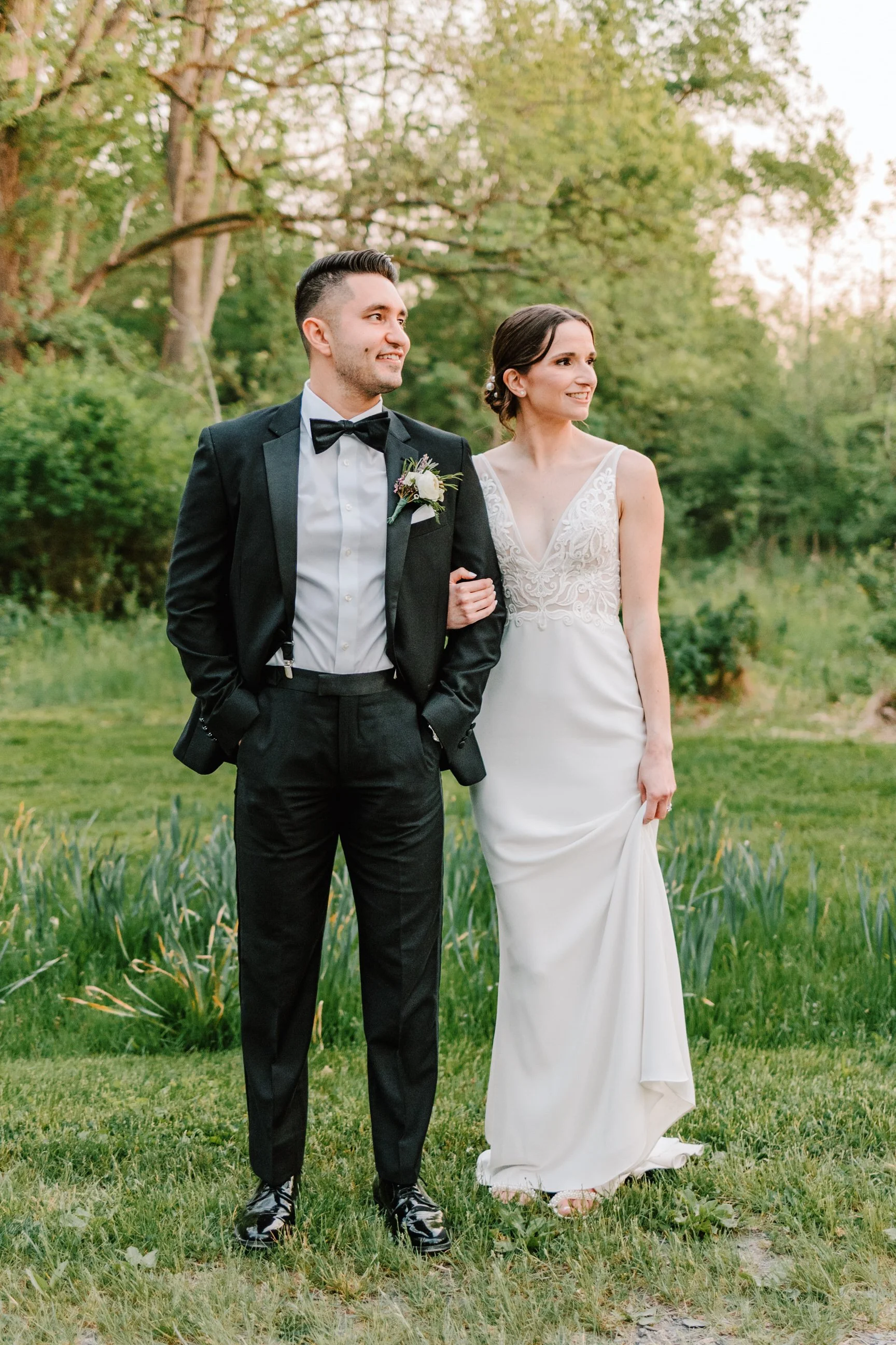 A bride and groom standing outdoors in a grassy area with trees in the background, smiling and holding arms, dressed in wedding attire.