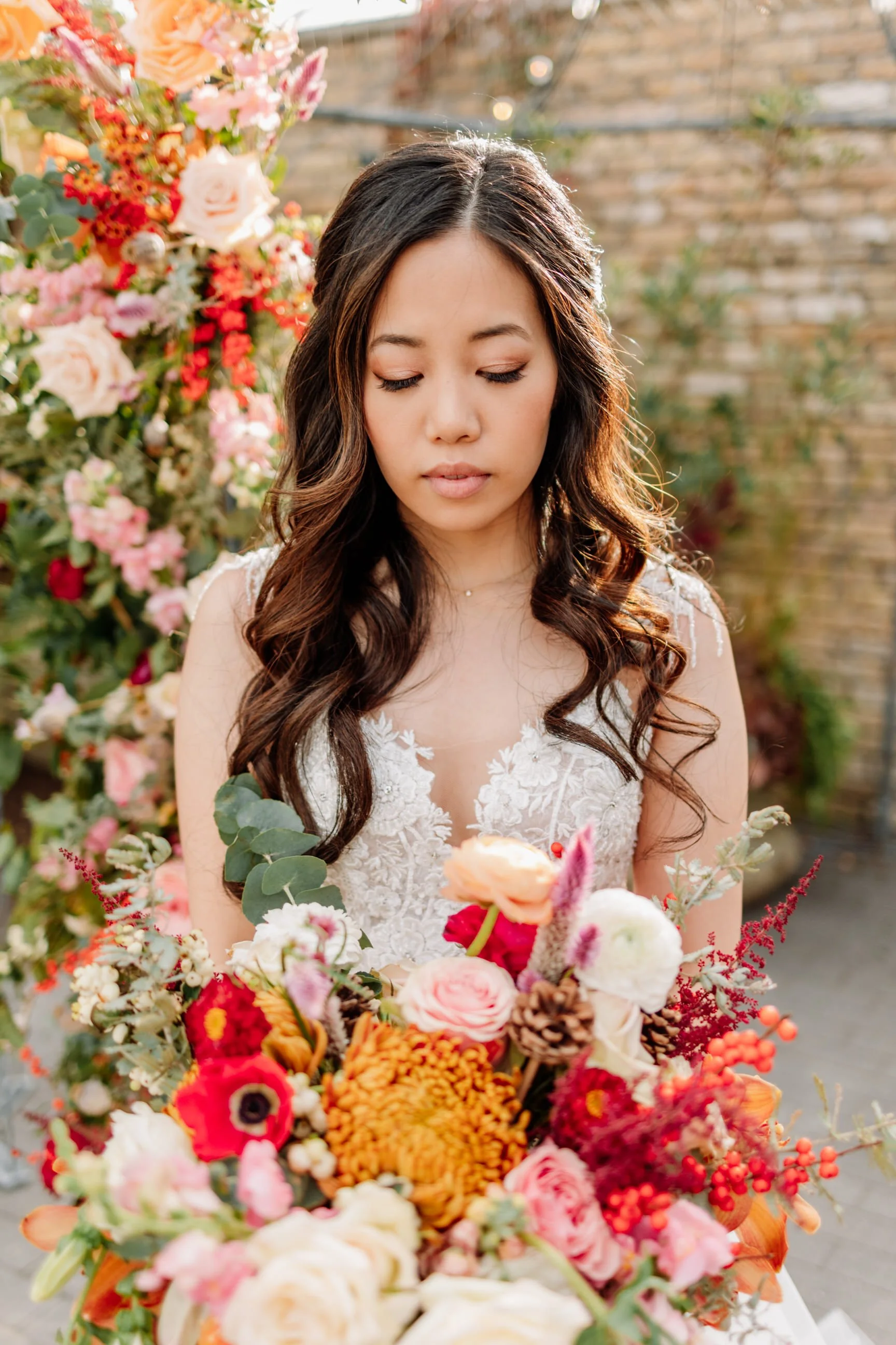 A woman with long wavy dark hair wearing a white lace dress standing outdoors with a colorful flower bouquet in front of her, and a background of a flowering bush and brick wall. Philadelphia wedding photographer