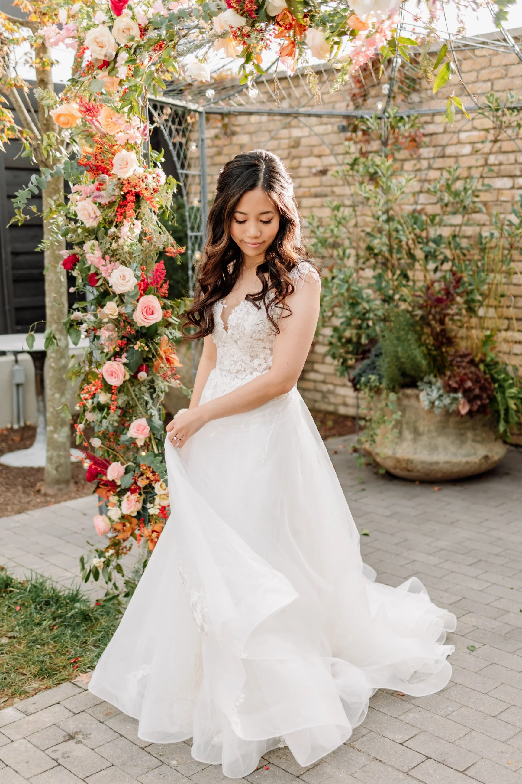 A woman in a wedding dress standing outdoors near a floral arch and potted plants.