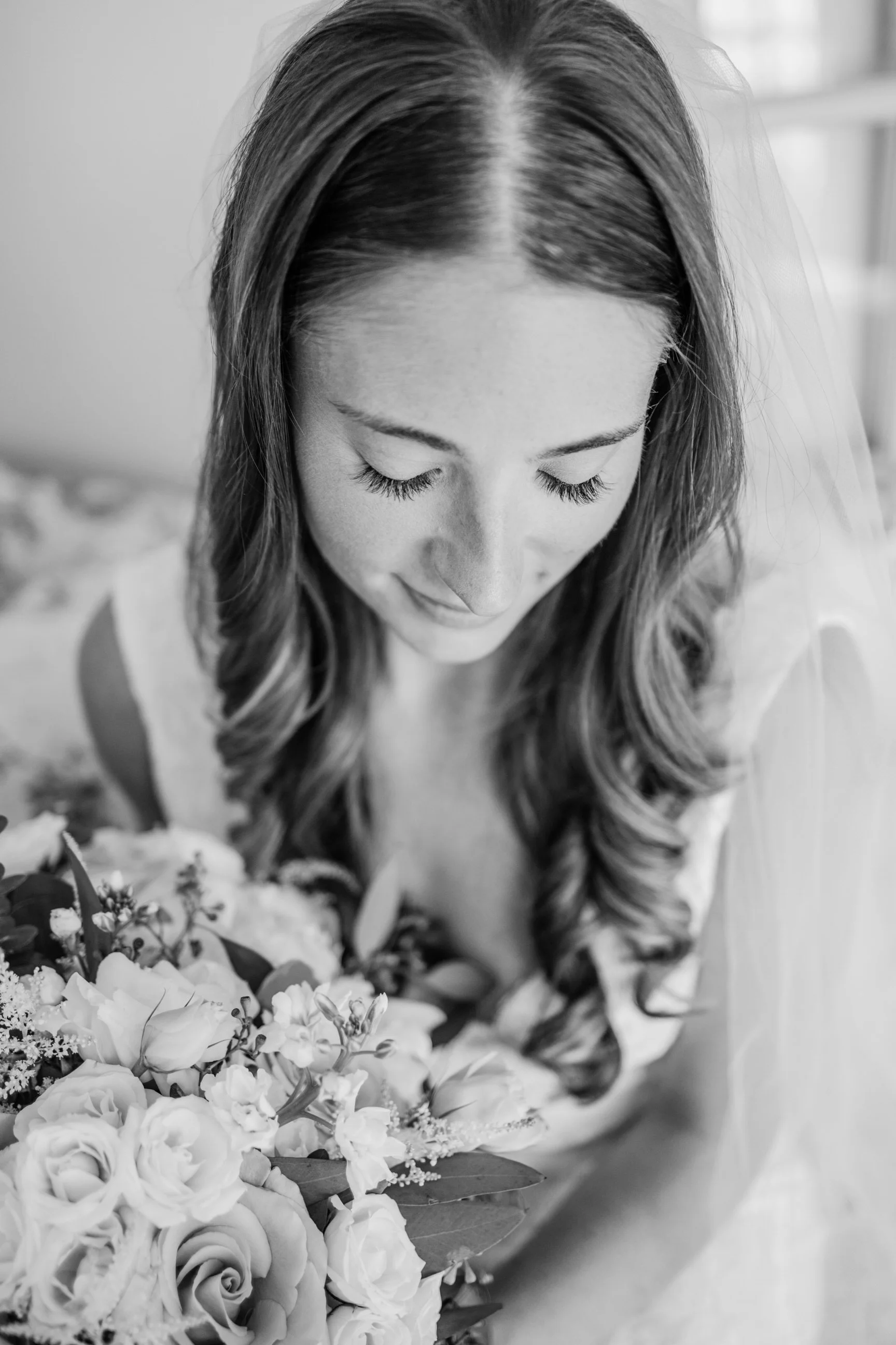 A bride with long wavy hair, wearing a veil and a wedding dress, looking down at a bouquet of flowers.
