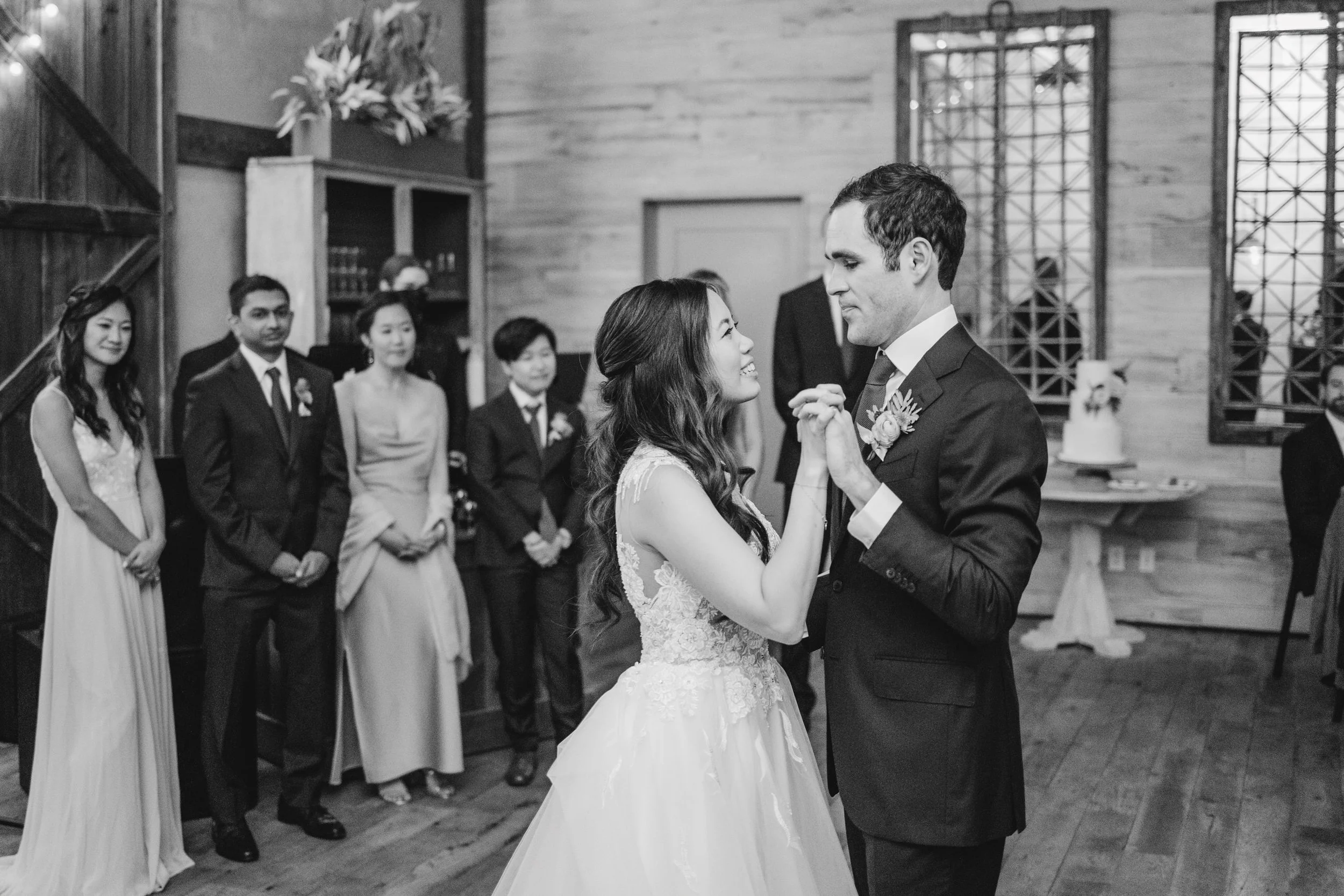 A bride and groom are dancing at their wedding reception, surrounded by guests watching. The bride is wearing a lace wedding gown, and the groom is in a dark suit with a boutonnière. The scene takes place indoors with wooden walls and a decorative la