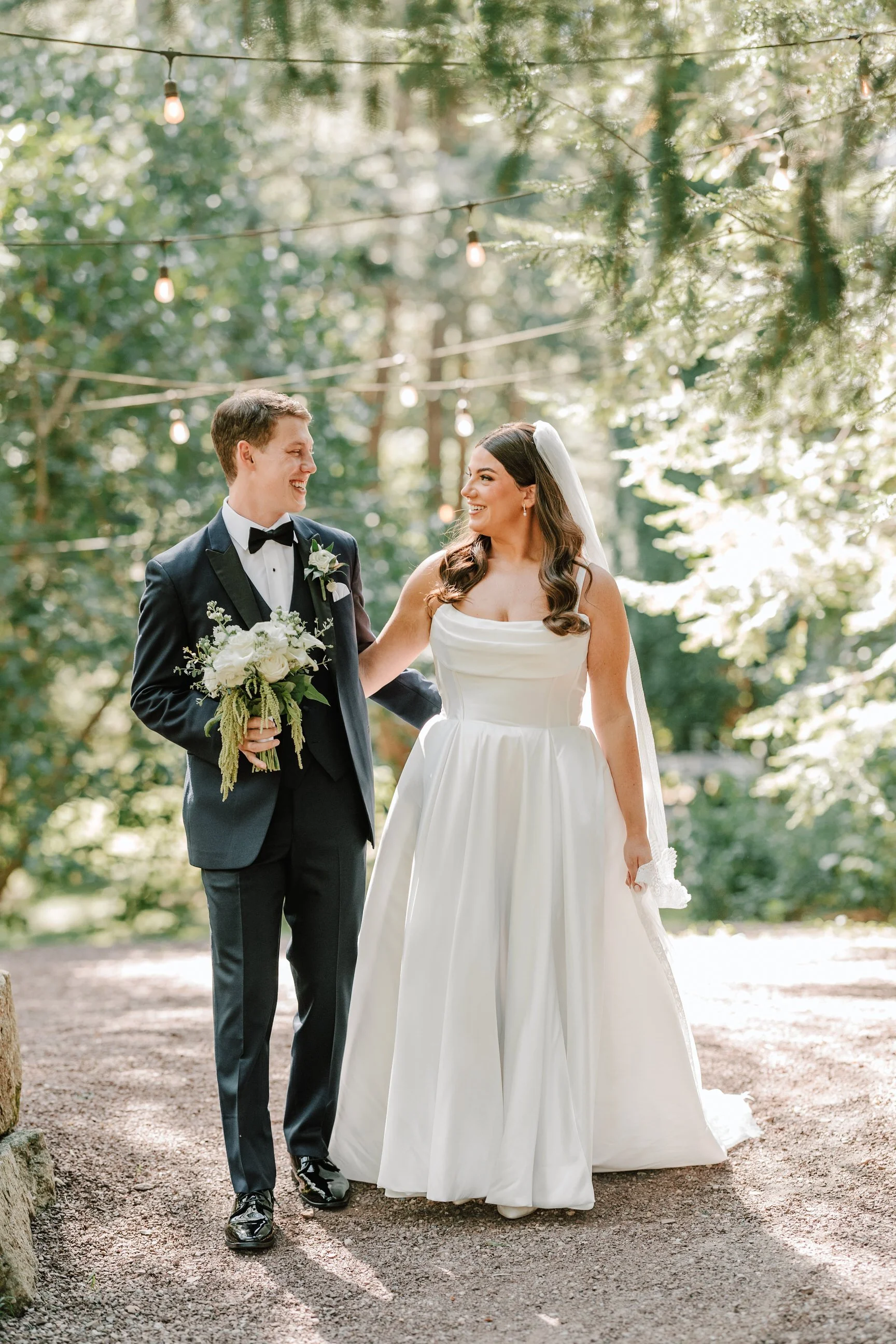 A bride and groom smiling at each other outdoors, with string lights hanging overhead, surrounded by trees, during a wedding ceremony.