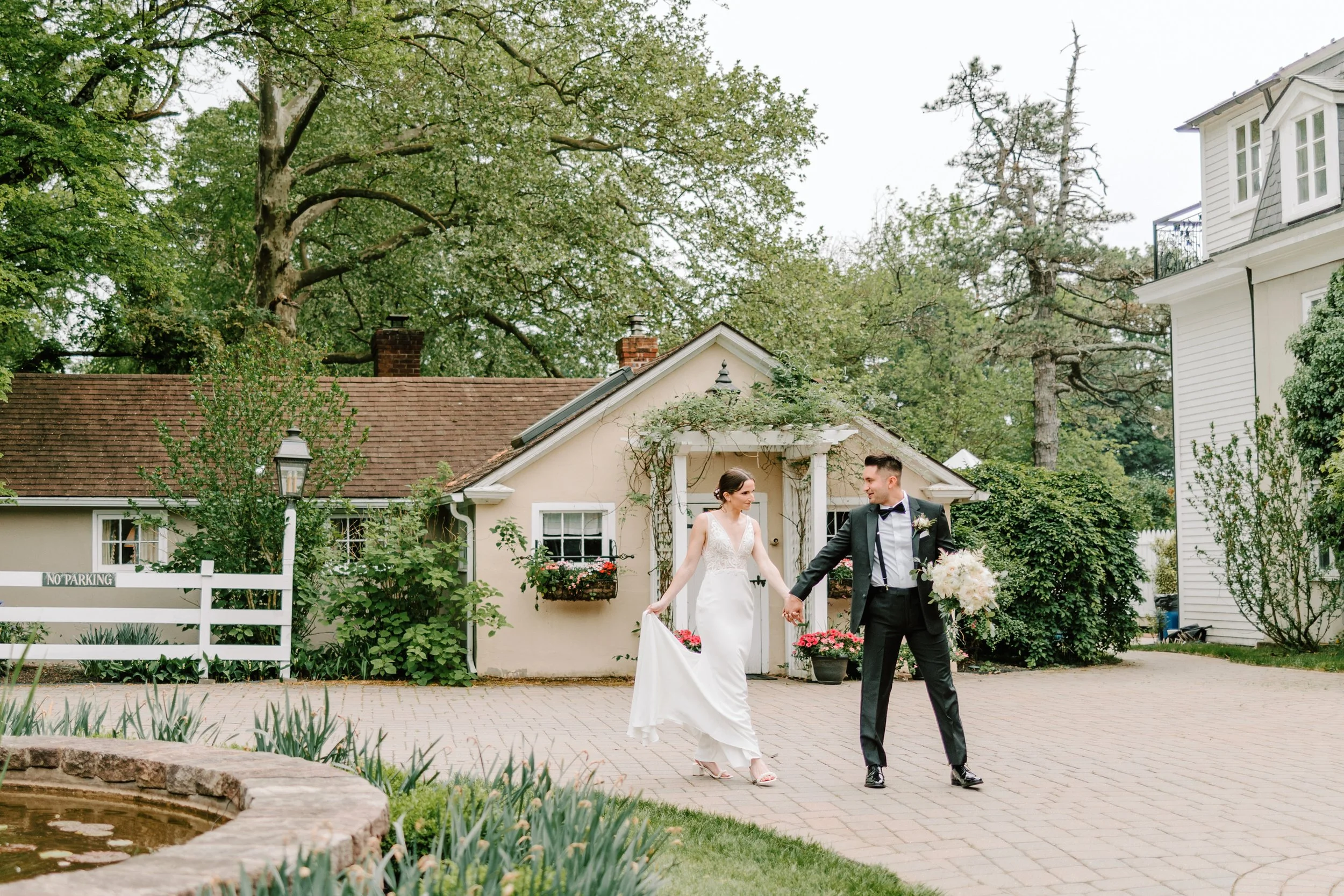 A bride and groom holding hands and walking outdoors near a garden and small house, celebrating their wedding day.