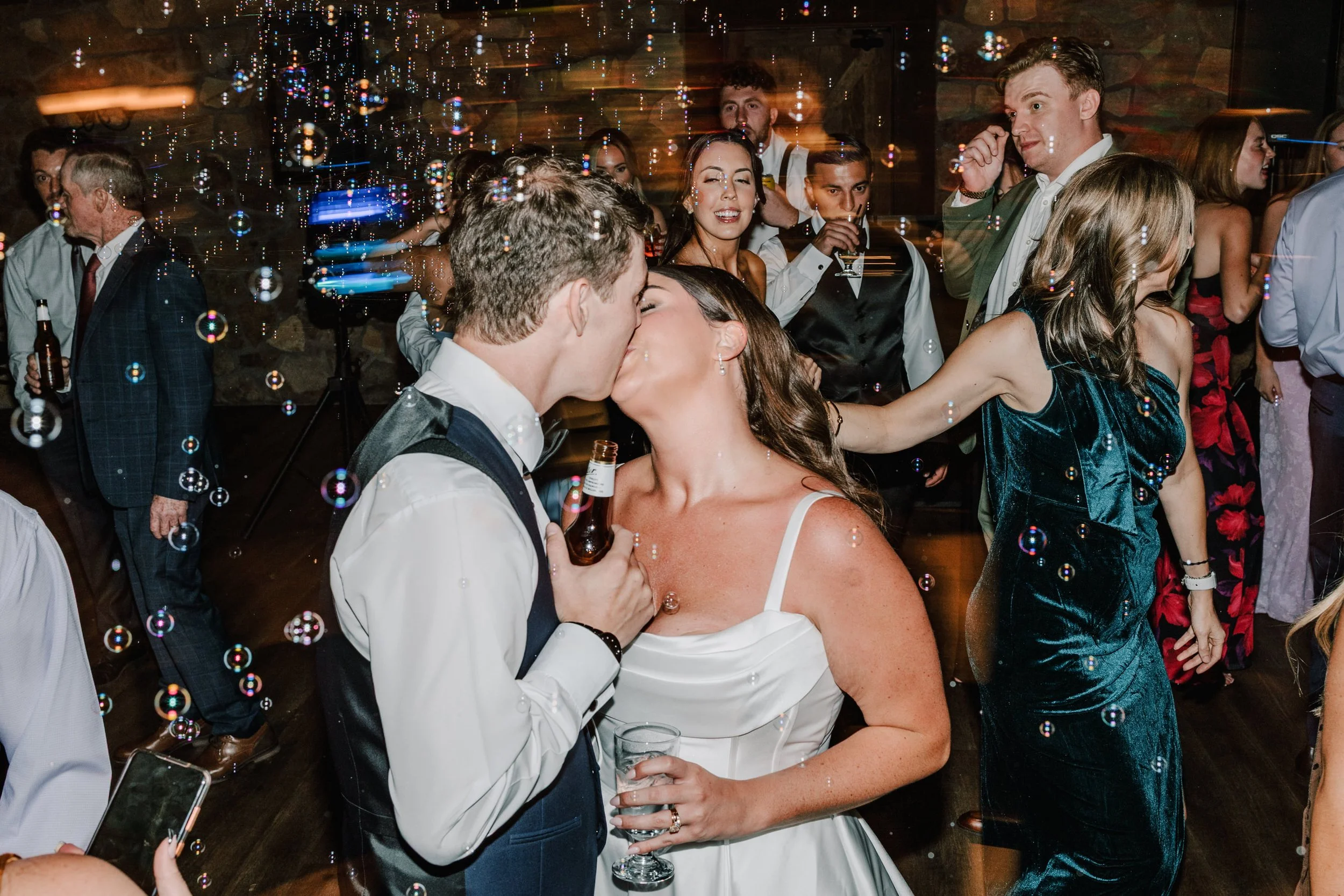 A wedding reception with a bride and groom sharing a kiss among guests, some surrounded by bubbles, in a warmly lit venue with wooden walls and dancing guests.