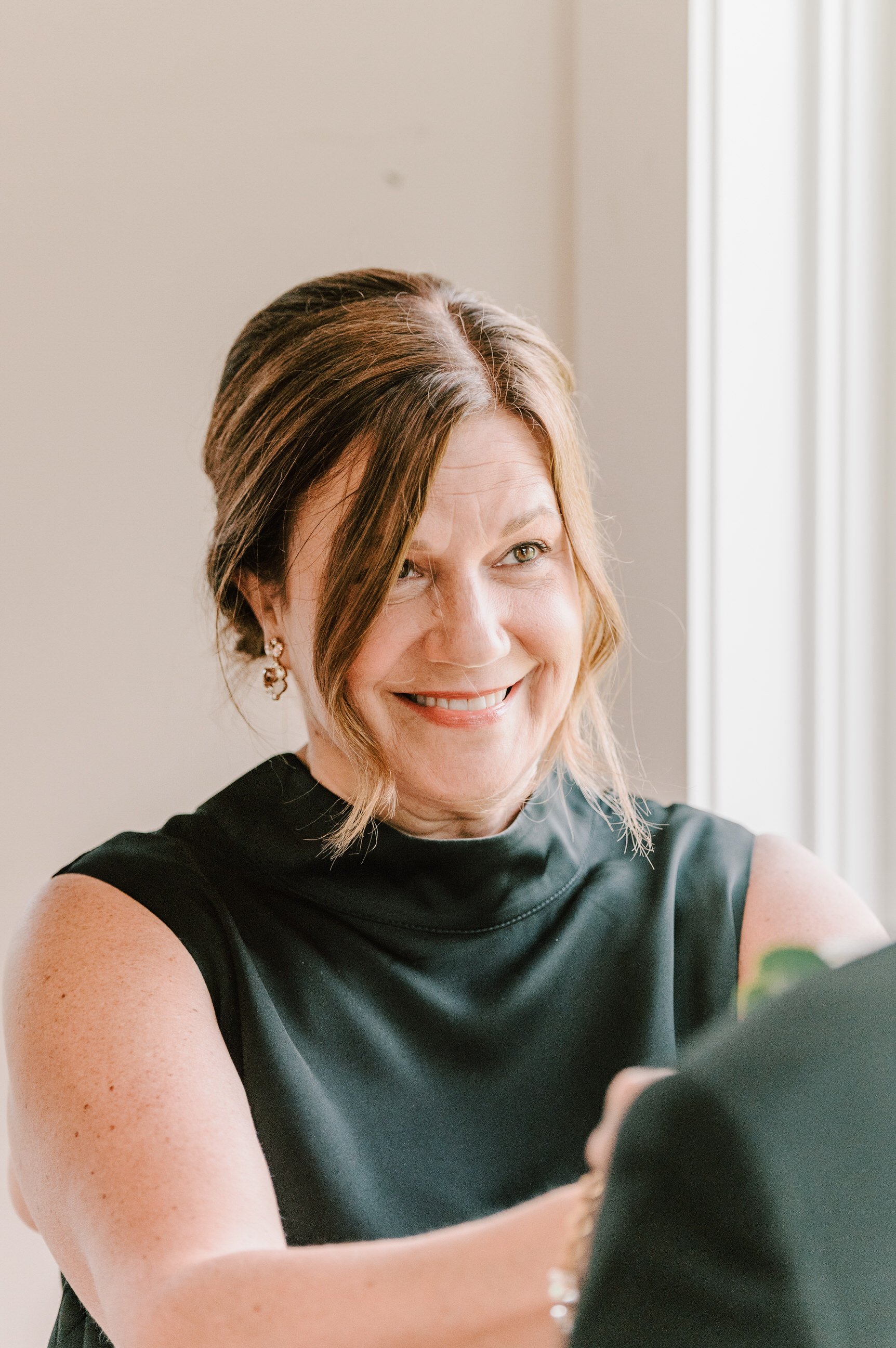 Smiling woman with short brown hair wearing a black sleeveless top, earrings, and a bracelet in front of a window with natural light.