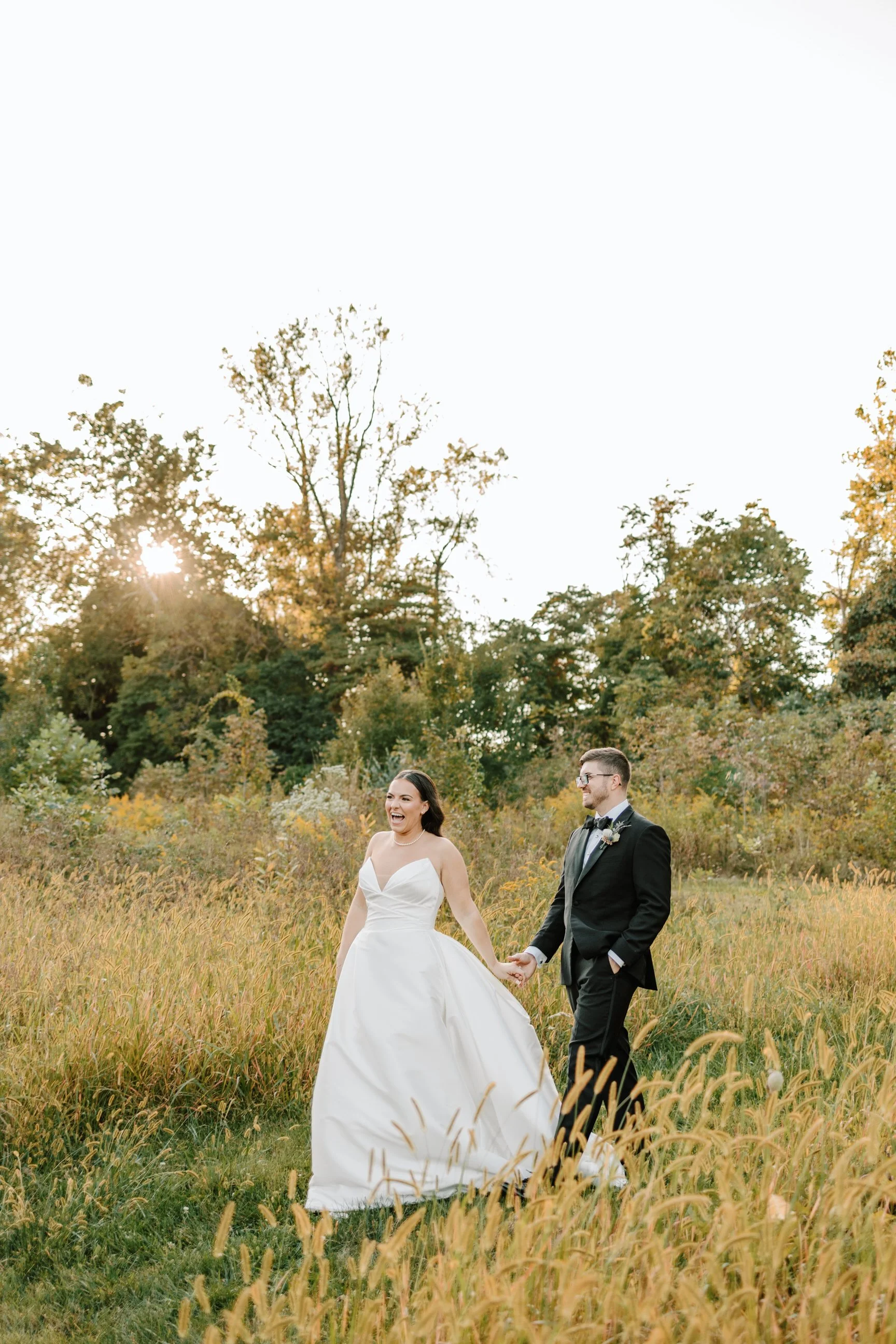 A bride and groom walking hand in hand through a grassy field at sunset, smiling and laughing. The bride wears a strapless white wedding gown and the groom is in a black tuxedo.