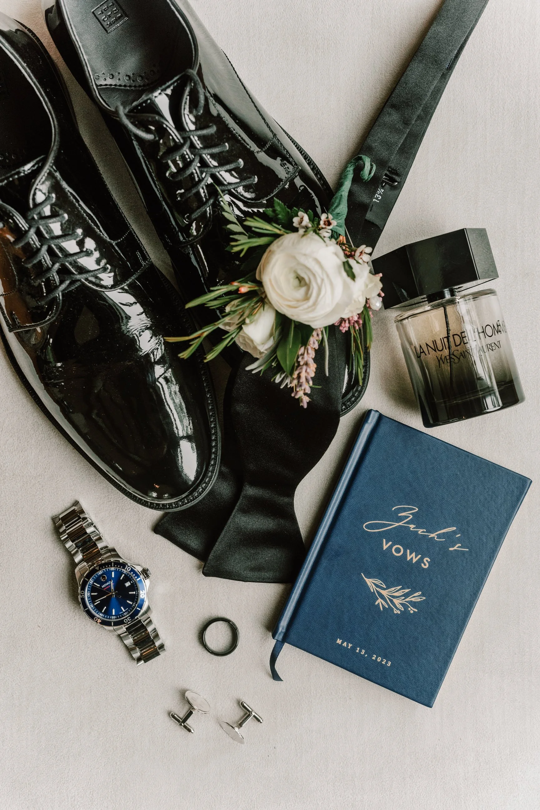 A flat lay of men's fashion and grooming accessories, including black patent leather dress shoes, a silver wristwatch with a blue face, a black leather belt, a black bowtie, a bouquet of white flowers with greenery, a black scented candle labeled 'La