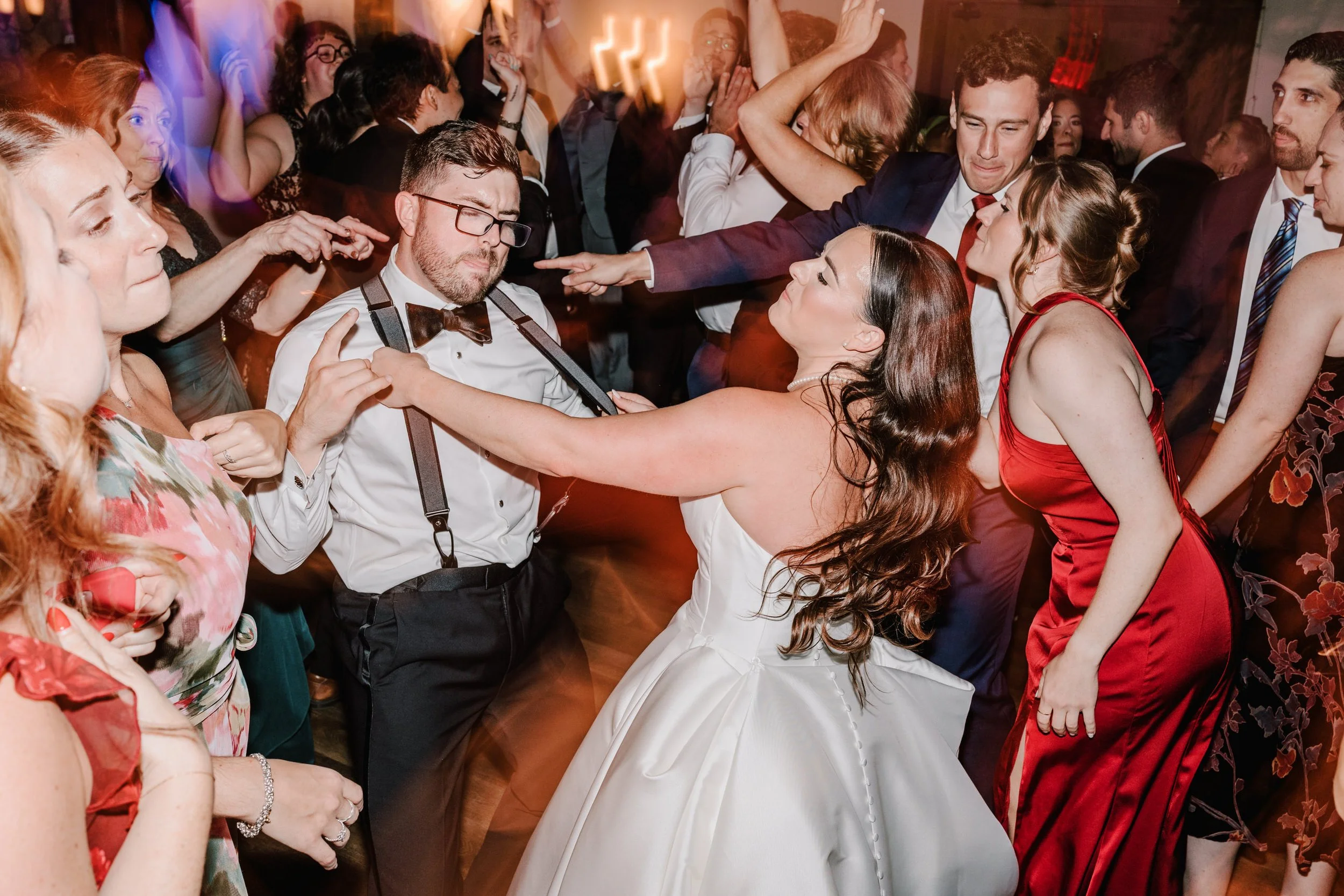 A bride and groom dance closely with guests at a wedding reception, surrounded by people in formal attire, some dancing and cheering.