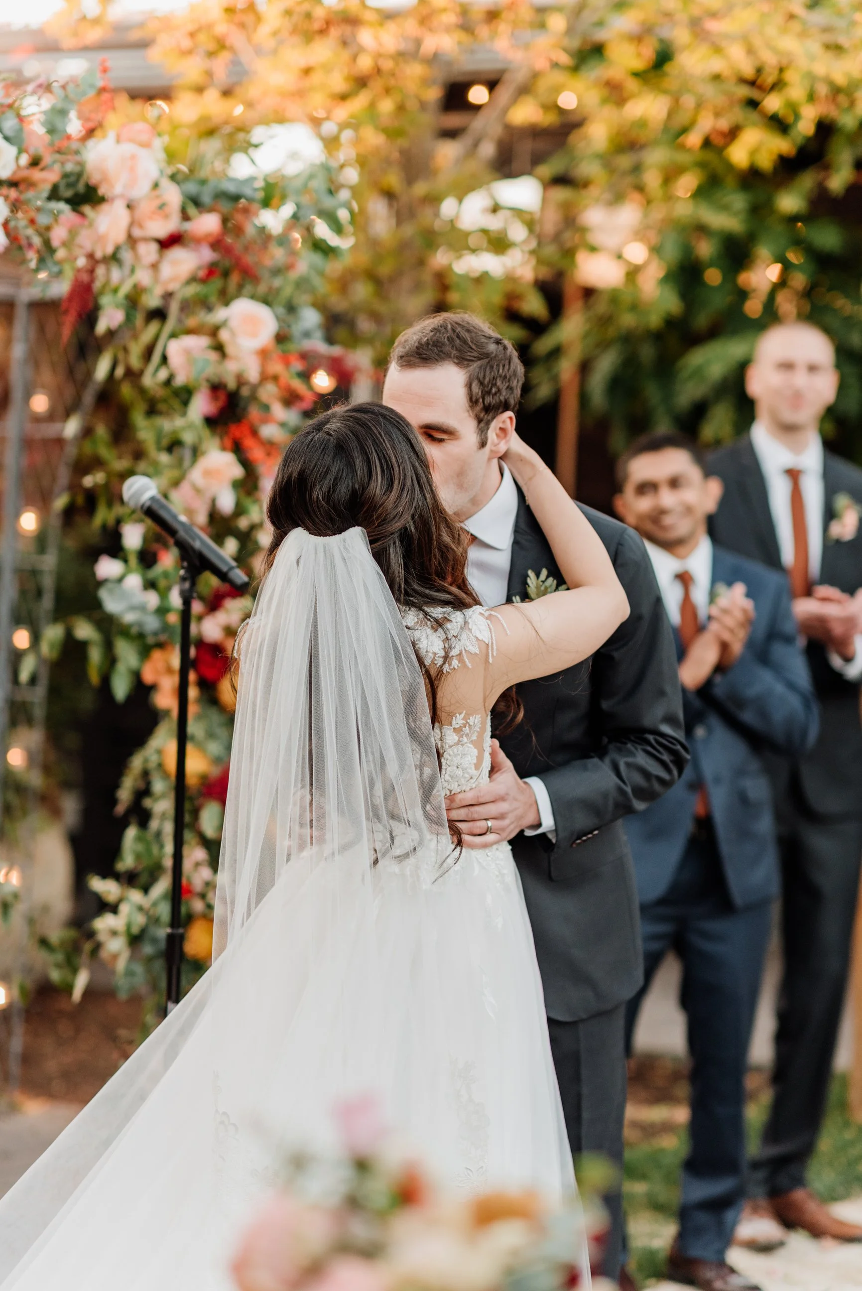 Bride and groom share a kiss at their outdoor wedding ceremony with two groomsmen clapping in the background, decorated with colorful flowers.