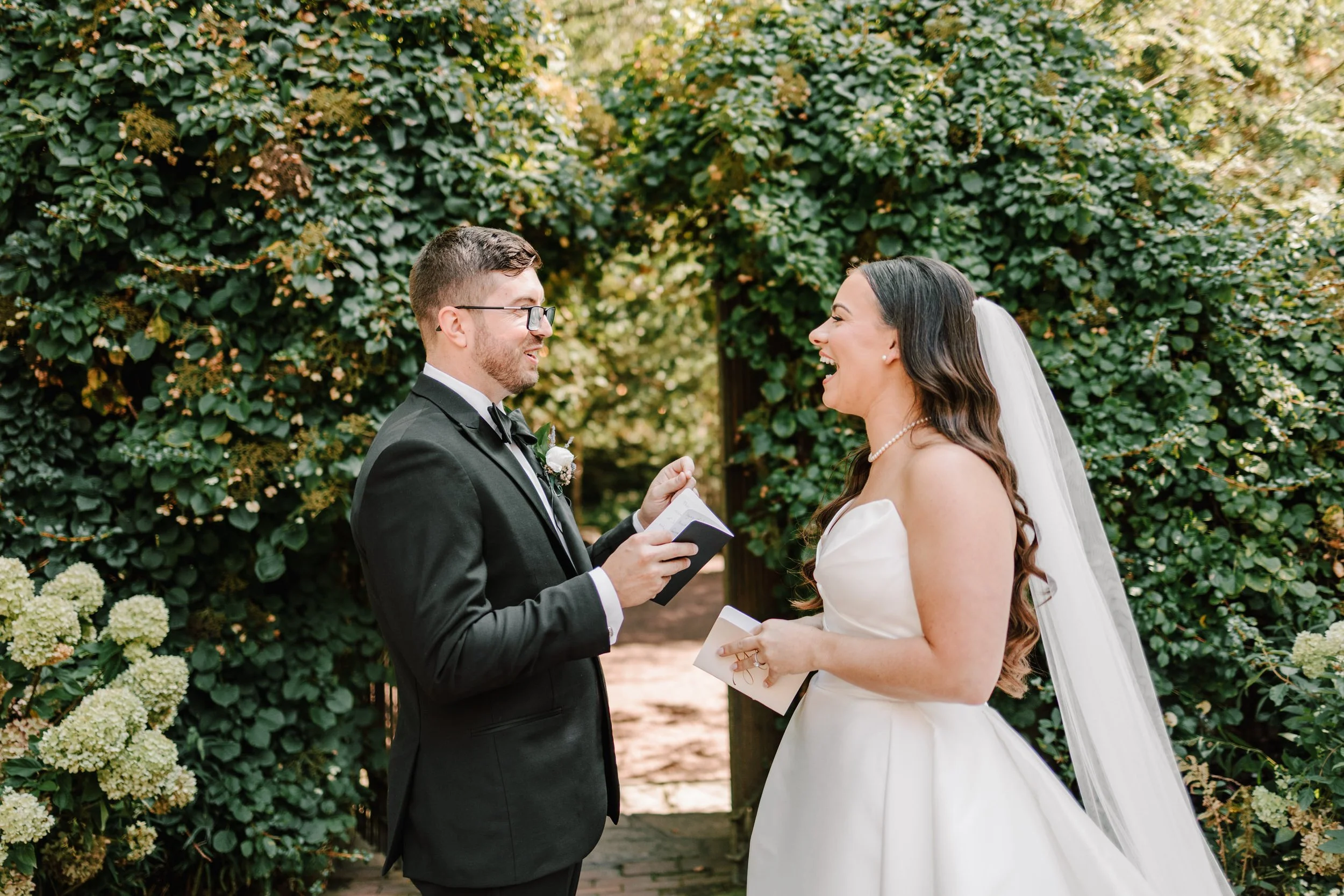 A bride and groom exchanging vows outdoors, with greenery and flowering plants in the background. The bride is in a strapless white wedding dress and veil, laughing, while the groom in a black tuxedo, glasses, and a boutonniere is holding a small not