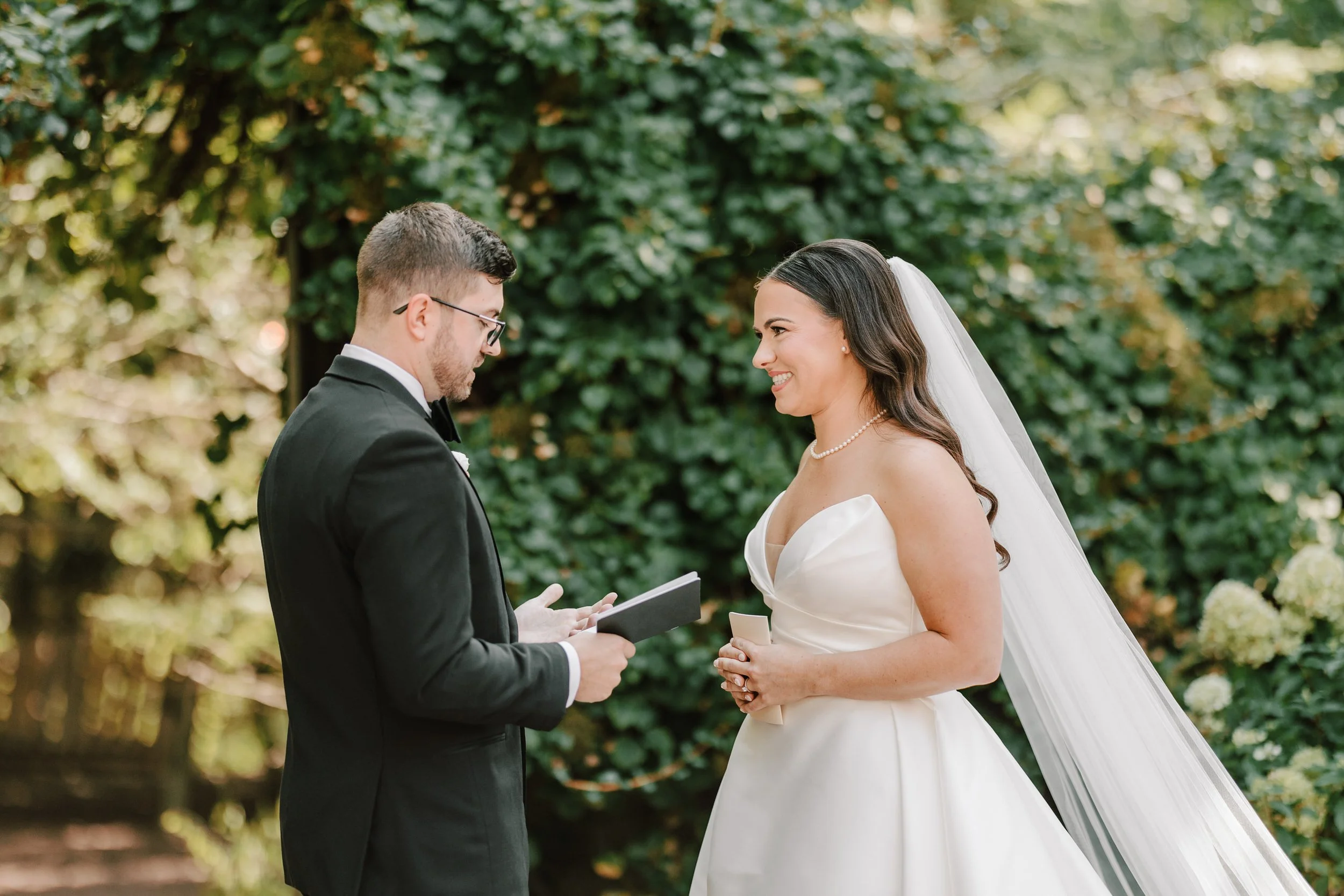 A bride and groom exchanging vows outdoors, with the bride smiling and wearing a white wedding gown and veil, and the groom holding a small book, dressed in a black tuxedo, surrounded by greenery.
