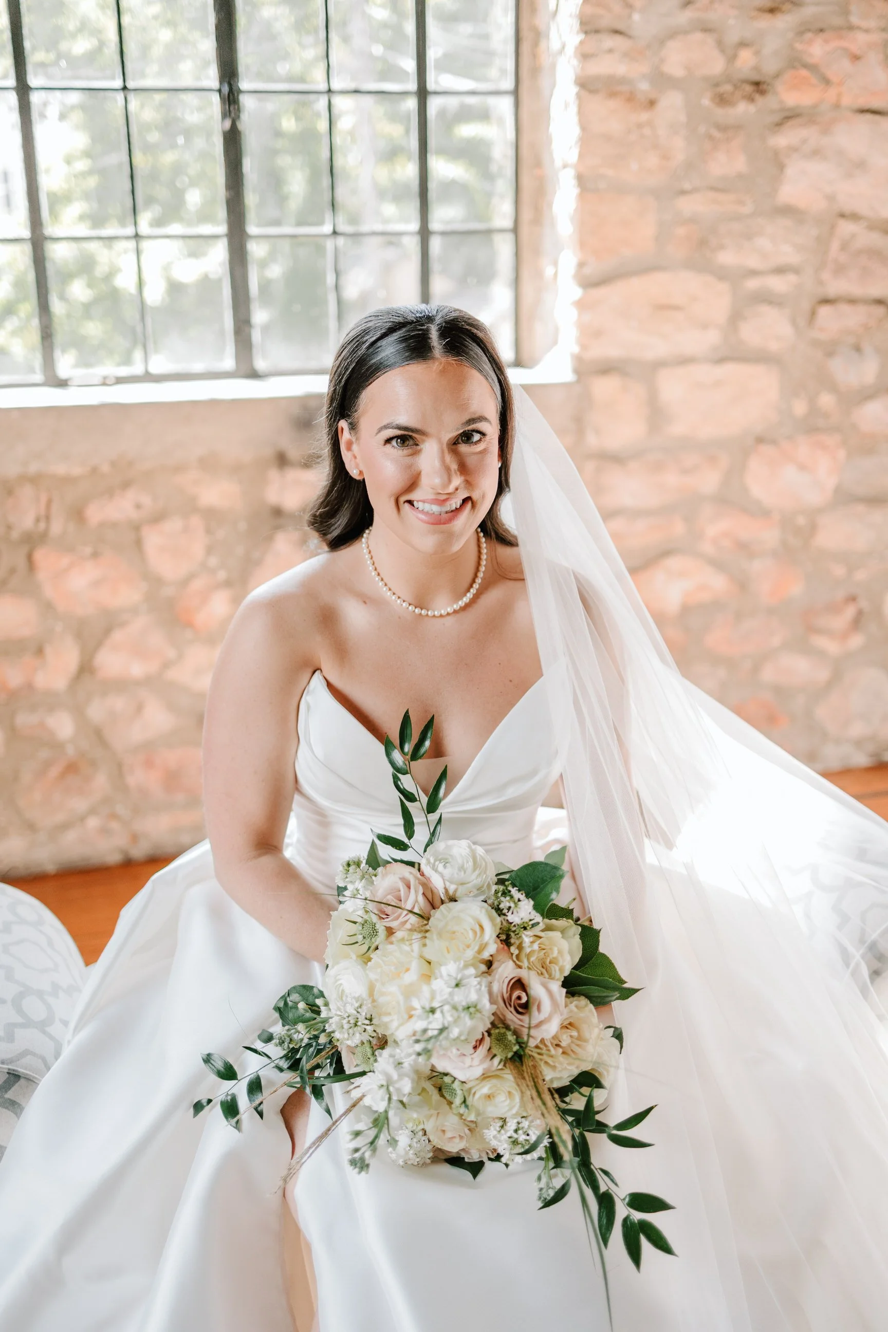 Bridal portrait of a smiling bride in a white wedding gown holding a bouquet of white and blush roses, posing indoors in front of a stone wall and a window.