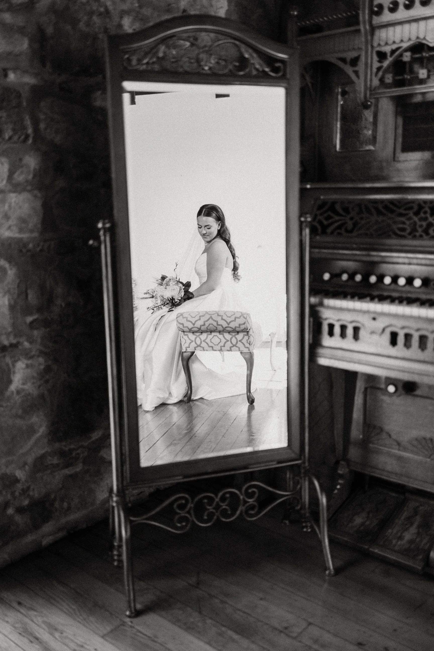 A woman in a wedding dress sitting on a bench, reflected in an ornate standing mirror, with a stone wall and musical instrument cabinet in the background.