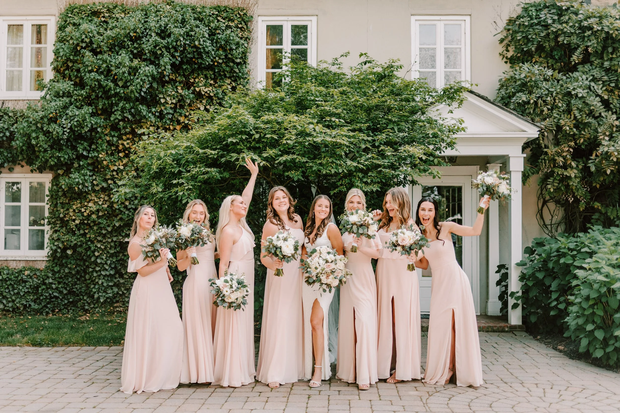 A group of eight women in matching light pink dresses standing outside in front of a house with lush green ivy and foliage, holding bouquets of white and pink flowers, smiling and celebrating.
