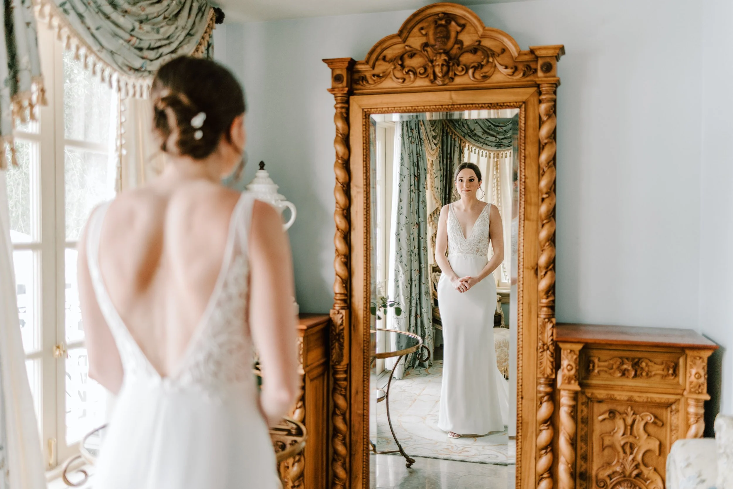 A bride in a white wedding dress standing in front of a large antique wooden mirror, looking at her reflection, with elegant curtains and vintage furniture in the background.