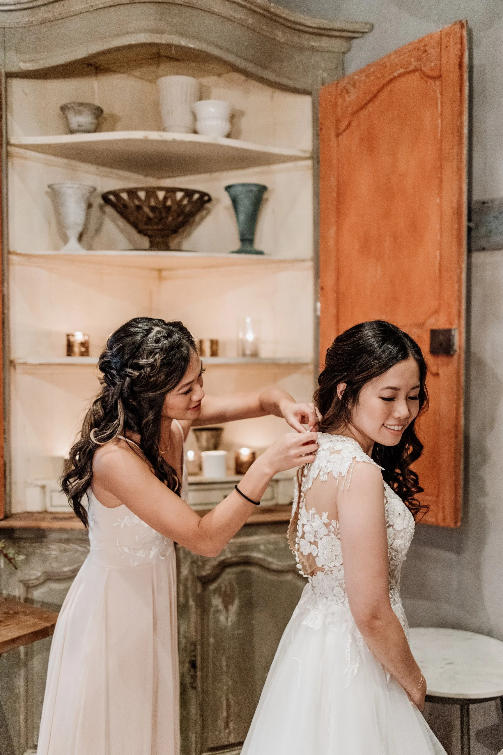 A woman in a white lace wedding dress smiling as another woman helps her fasten her dress near a rustic cabinet with decorative vases and candles.