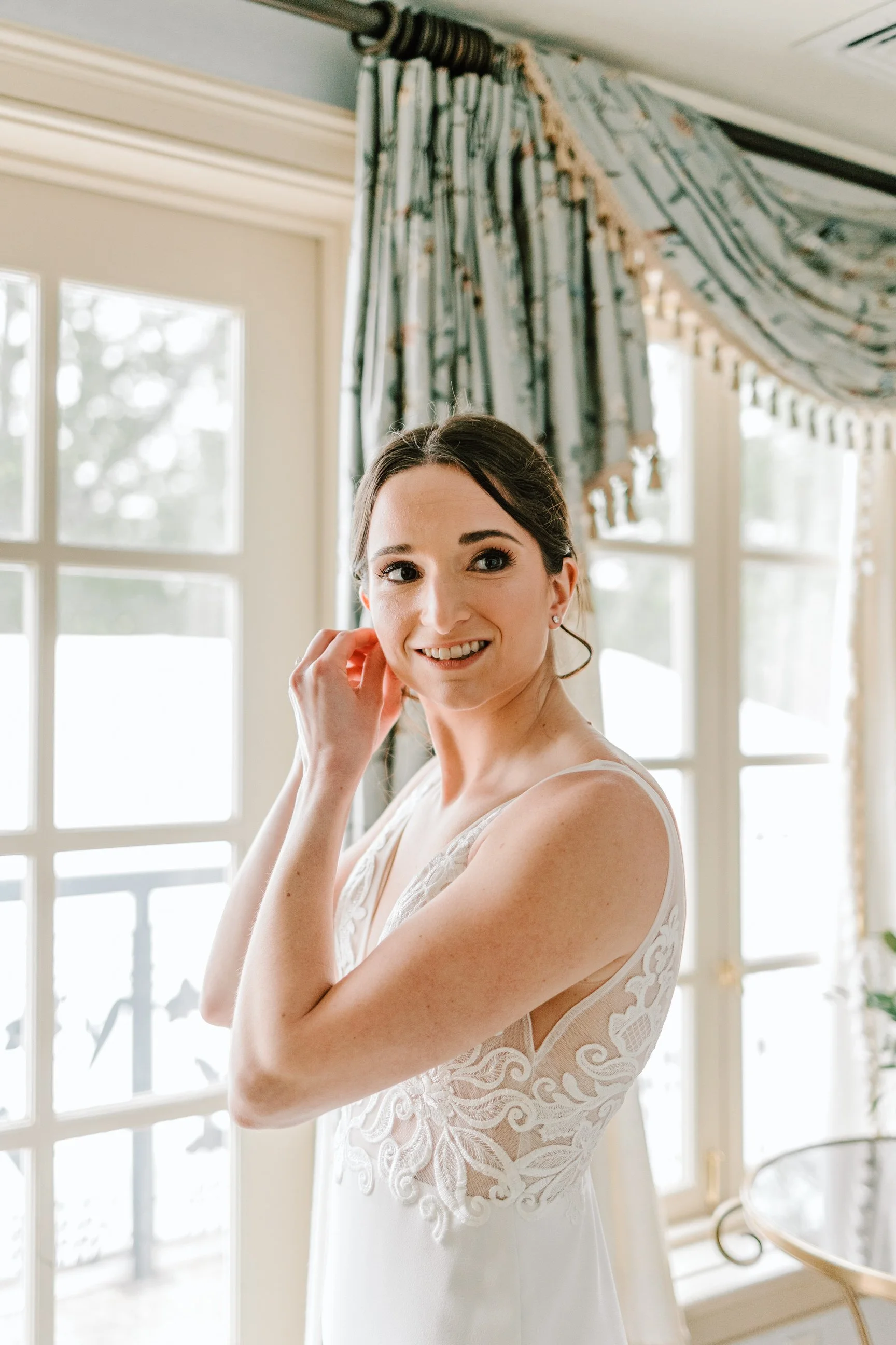 A woman in a white wedding dress standing indoors near large windows with floral curtains, smiling and touching her earring.