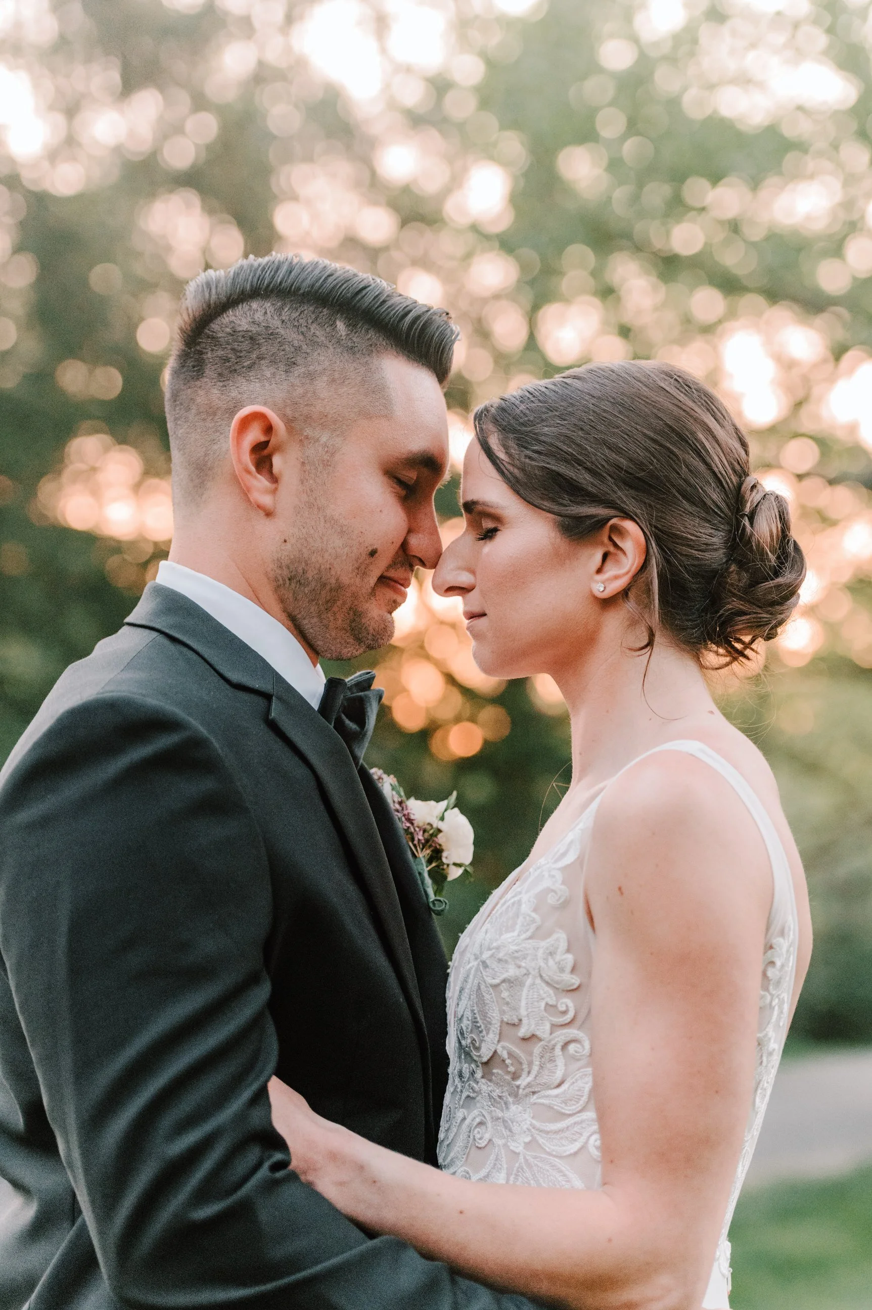 A newlywed couple with their foreheads touching, eyes closed, outdoors during sunset. The groom is in a black suit with a bow tie, and the bride is in a lace wedding dress, with her hair styled in an elegant updo.