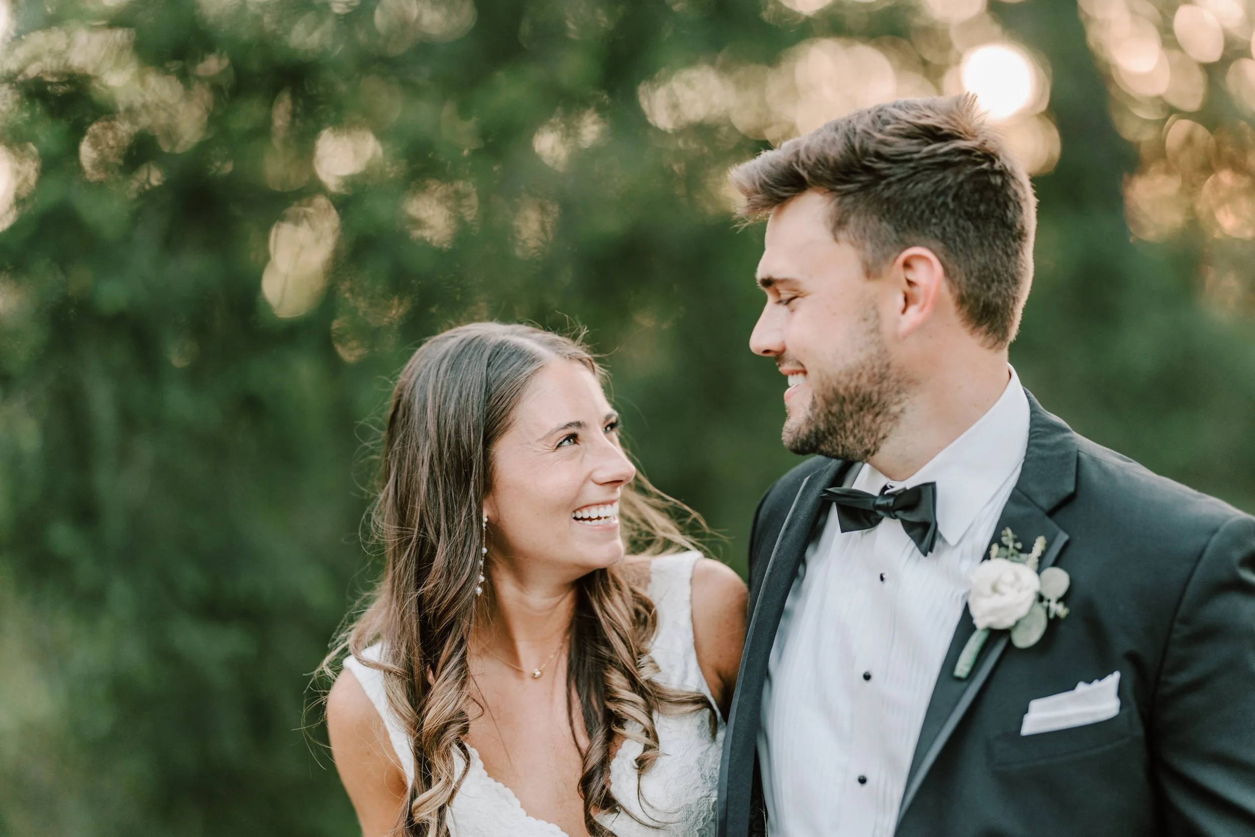 A bride and groom smiling at each other outdoors during sunset, with blurred greenery in the background.
