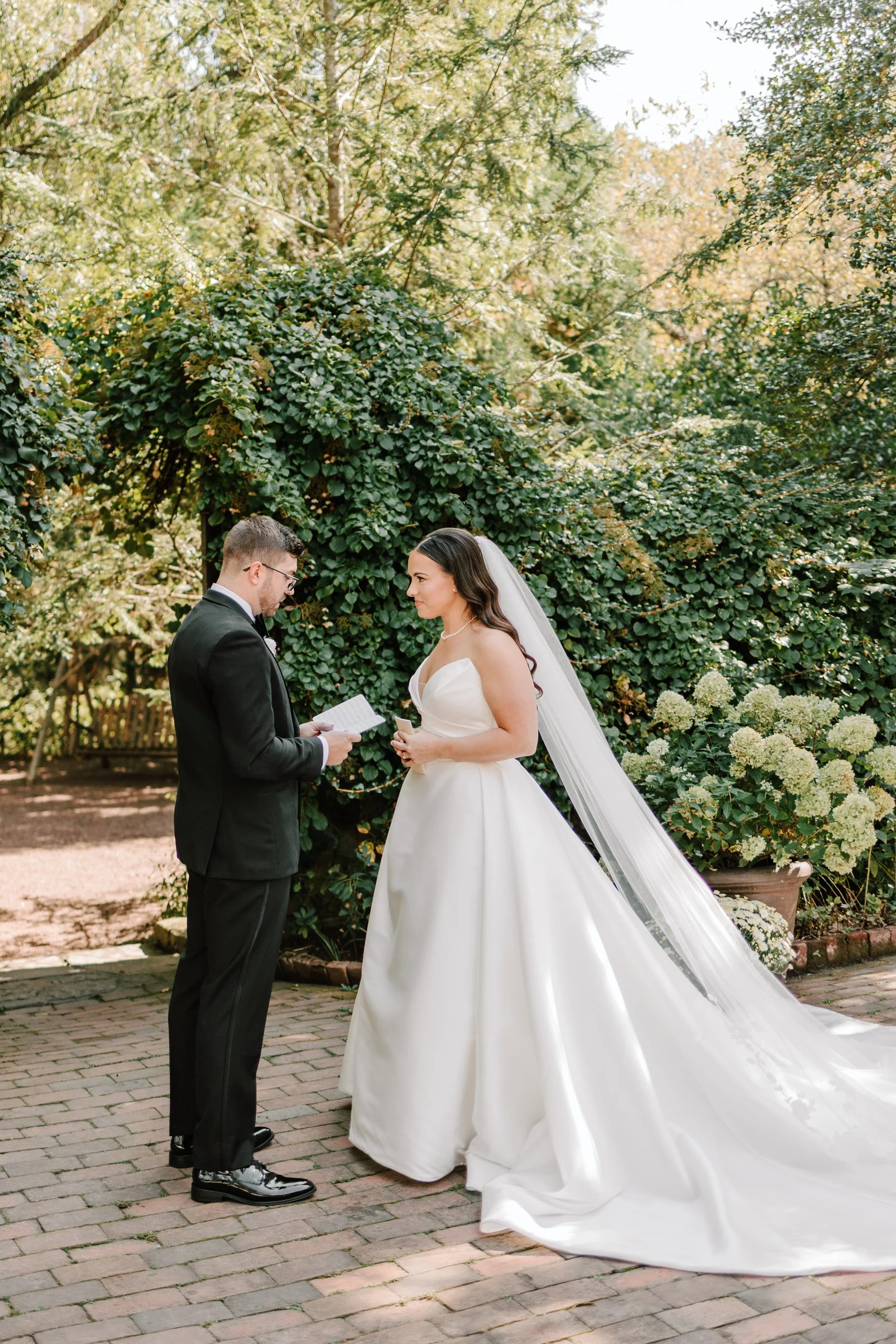 A bride and groom exchanging vows outdoors on a brick-paved area surrounded by greenery and flowering plants.