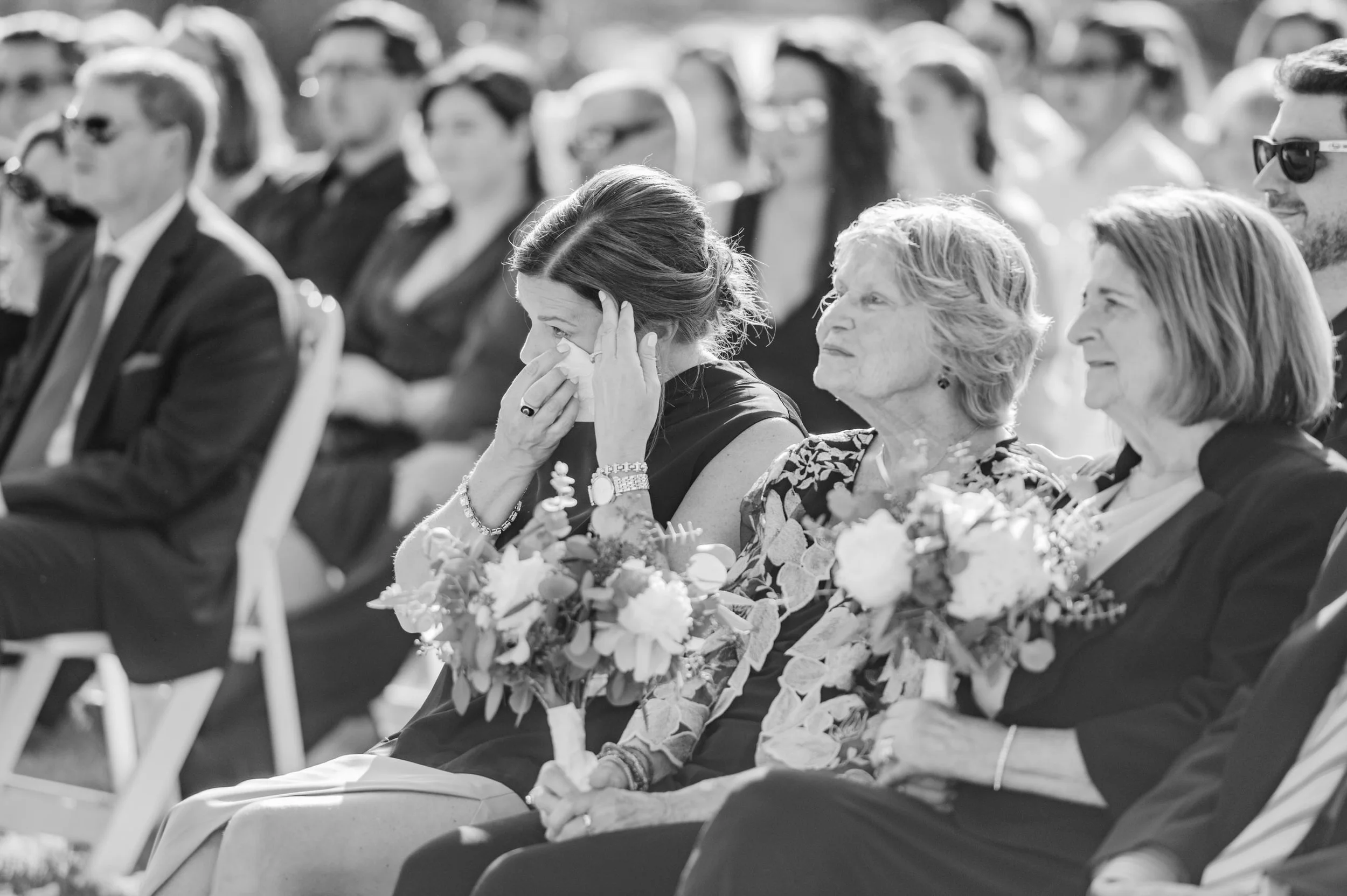 A group of women and men seated outdoors, attending a formal event. One woman in the middle is crying and wiping her eyes with a tissue, holding a bouquet of flowers. The others look solemn, and some women are also holding flowers.