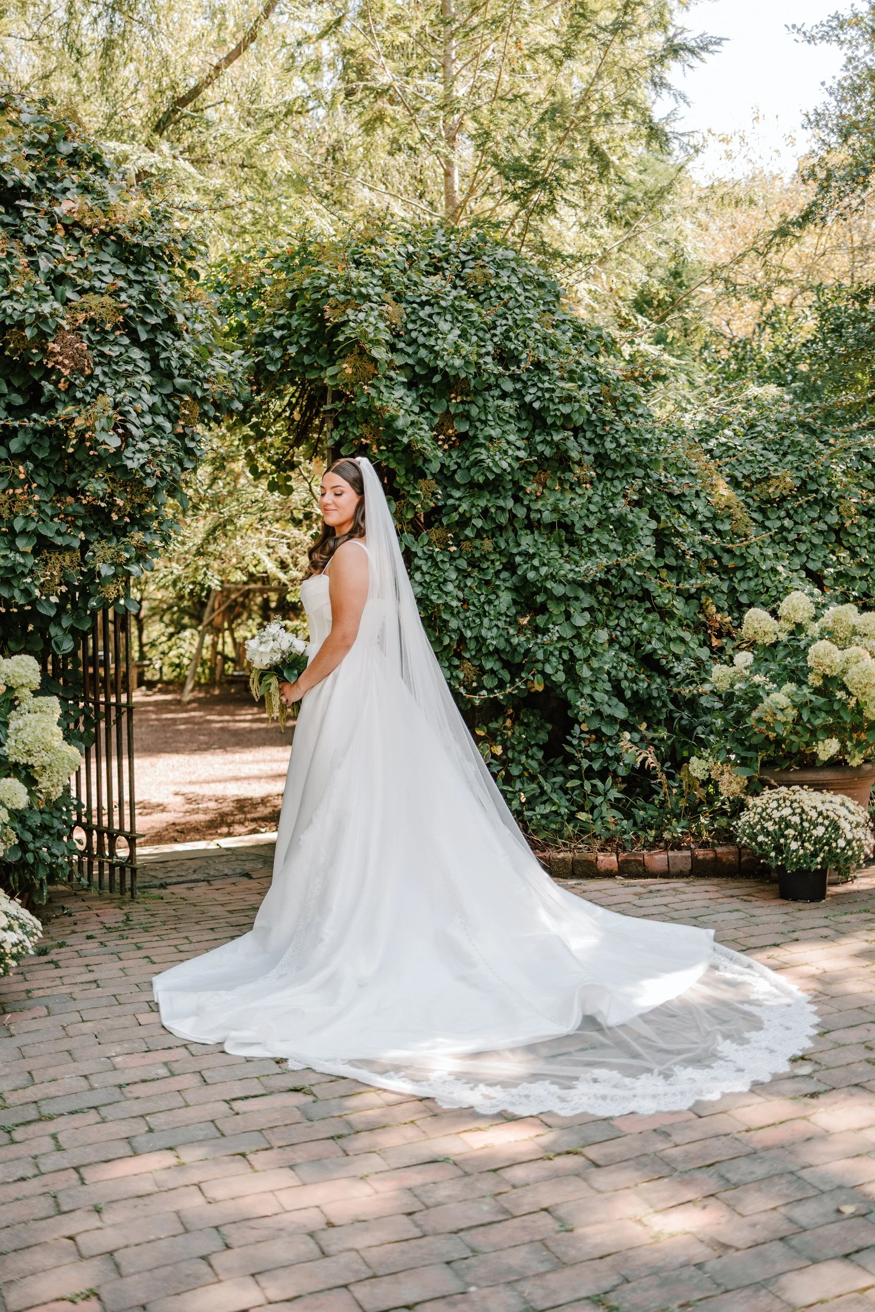 A bride standing outdoors on a brick pathway, holding a bouquet of white flowers. She is in a white wedding gown with a long train and a veil, surrounded by green foliage and flowers.