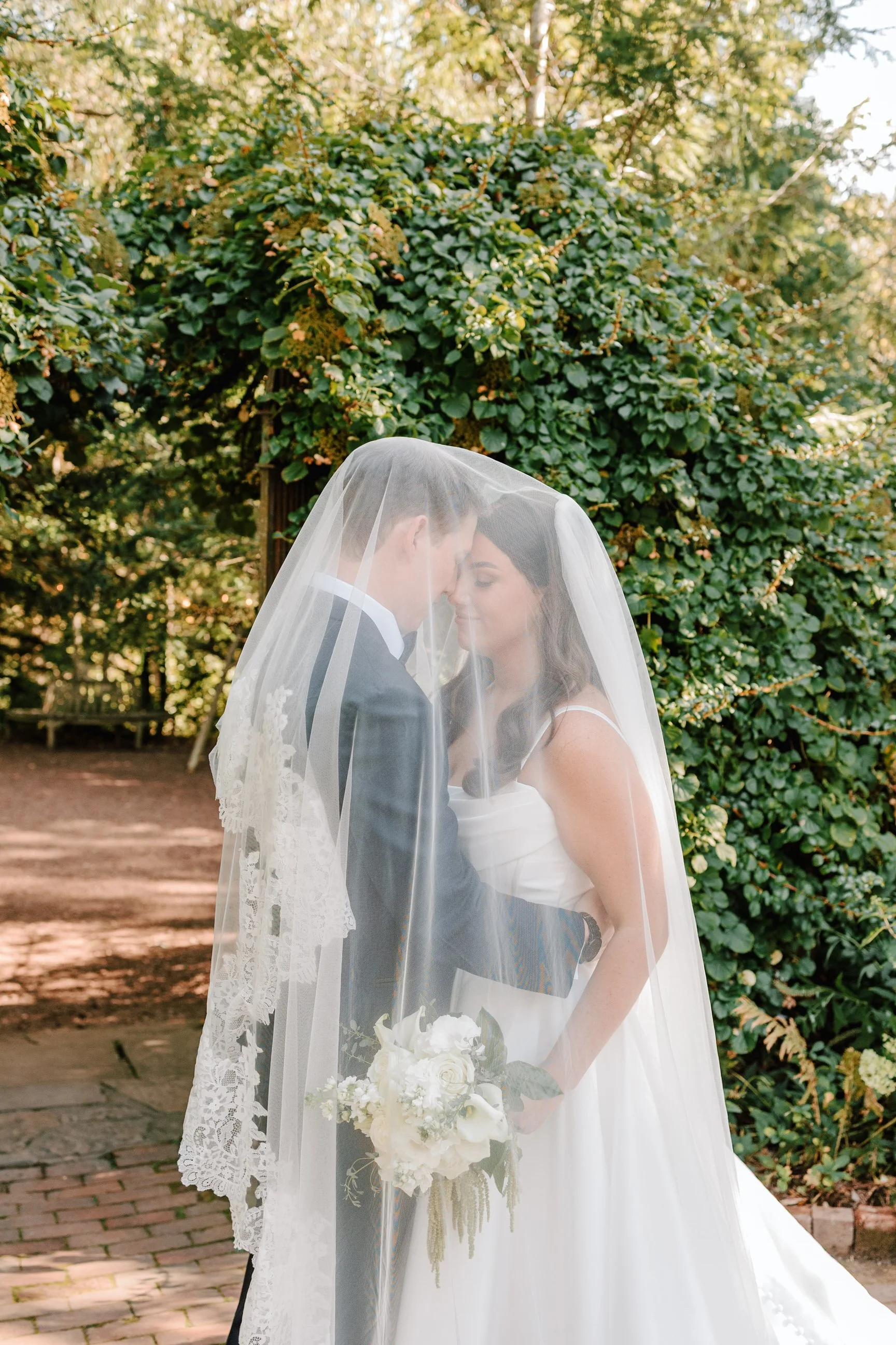 A bride and groom stand close together under a lace veil, touching foreheads, in an outdoor garden setting. The bride holds a bouquet of white flowers, and a leafy green backdrop surrounds them.
