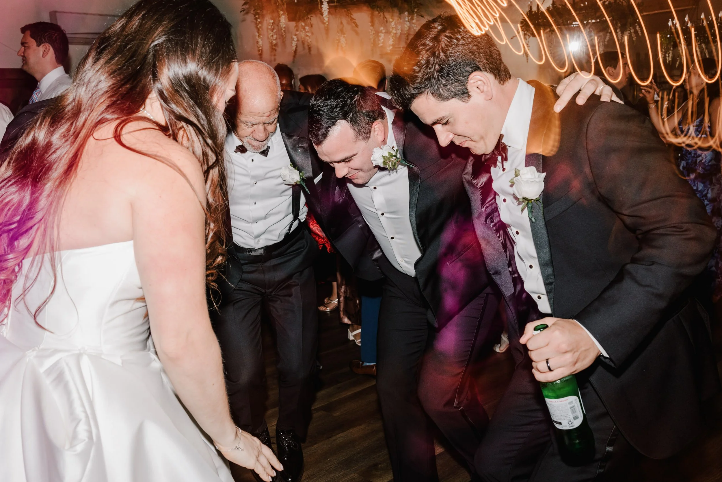 People dancing in tuxedos and a wedding dress at a wedding reception, with colorful lights and a festive atmosphere.