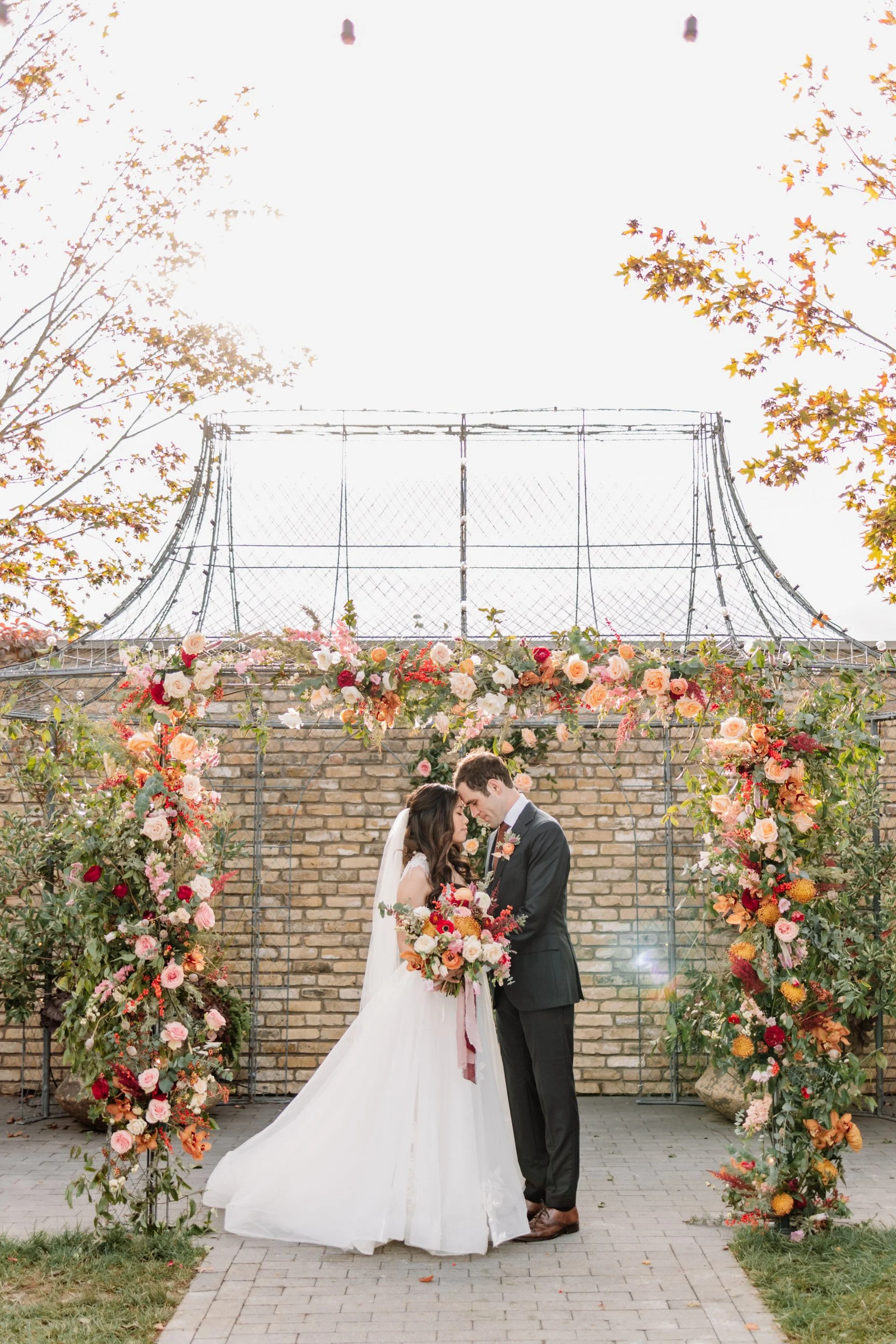 A bride and groom face each other with foreheads touching during their wedding ceremony, standing under a floral archway on a paved pathway near a brick wall, with trees on either side.