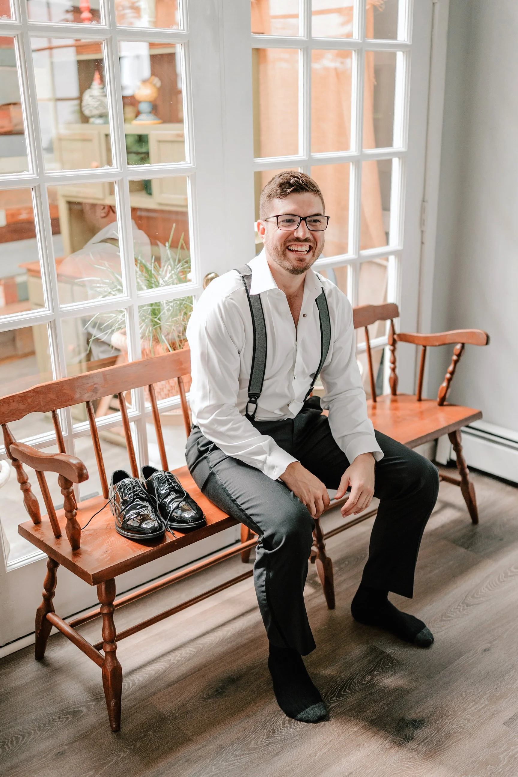 A man with glasses, wearing a white shirt, suspenders, and black pants, sitting on a wooden bench, smiling, with black polished shoes placed beside him.