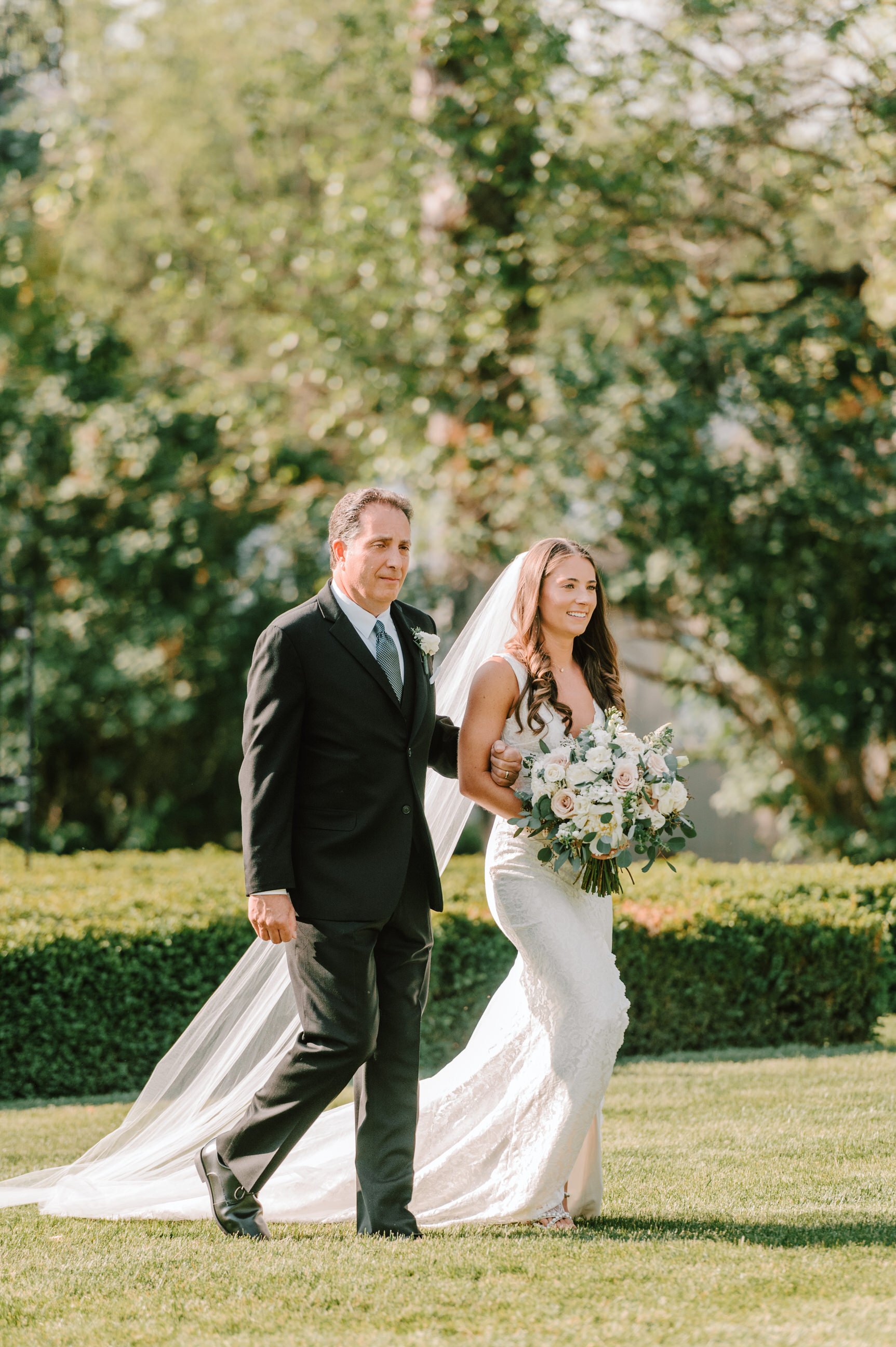 A bride walking outdoors on her wedding day, being escorted by a man in a black suit. The bride is in a white wedding dress holding a bouquet of white and pink roses, with a veil and long brown hair. The background features green trees and sunlight.