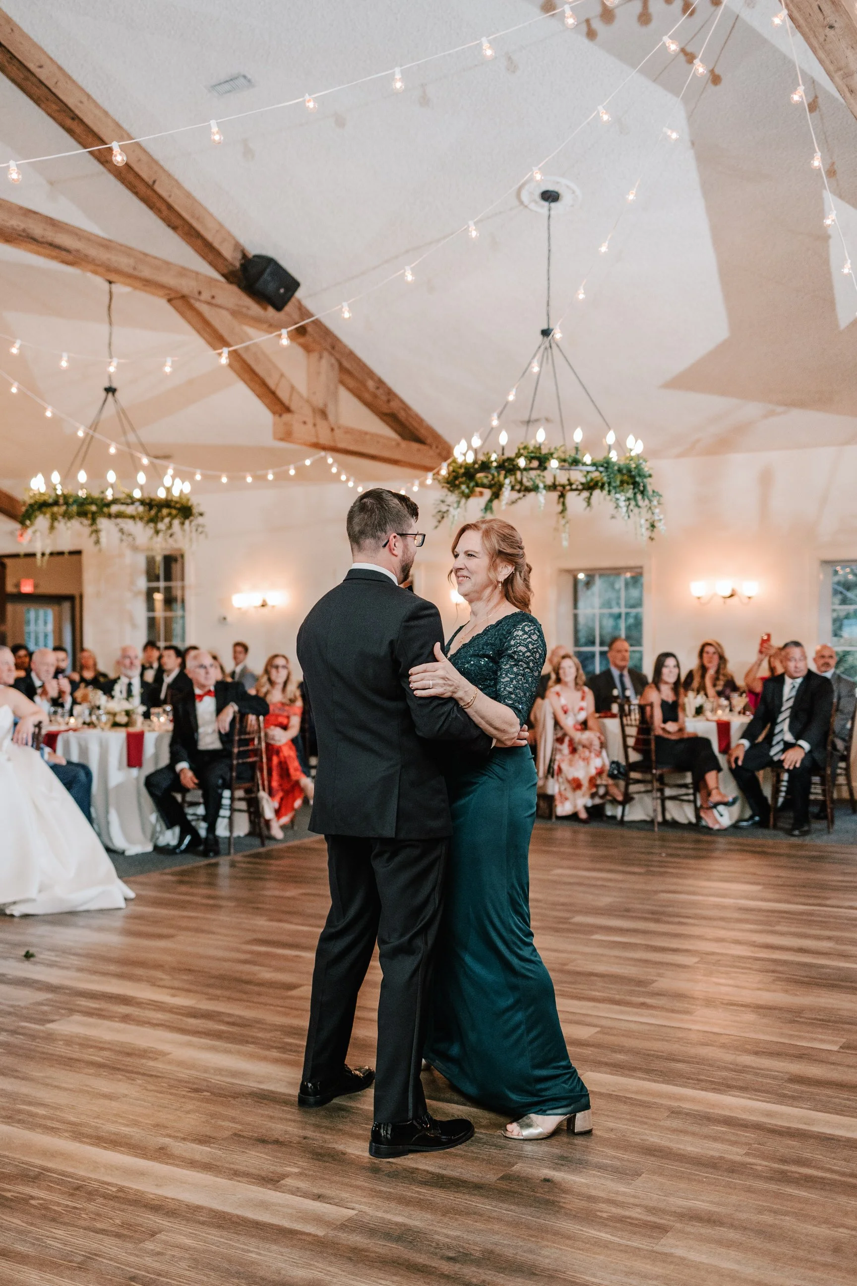 A mother and son dancing at a wedding reception, with guests sitting at tables in the background.